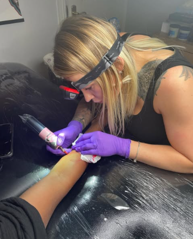 A tattoo artist wearing purple gloves and a headlamp ink an arm tattoo in a tattoo studio.