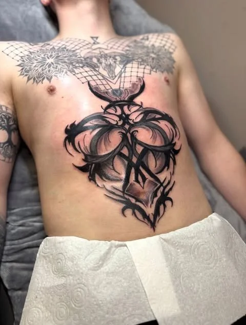 Close-up of a person's torso showing multiple tattoos, including a black and gray floral design on the chest, an intricate heart with tribal elements on the stomach, and a small tattoo near the shoulder.