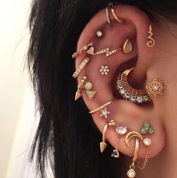 Close-up of a person's ear adorned with multiple earrings, including gold, diamond, and gemstone designs, featuring hoops, studs, and dangling pieces.