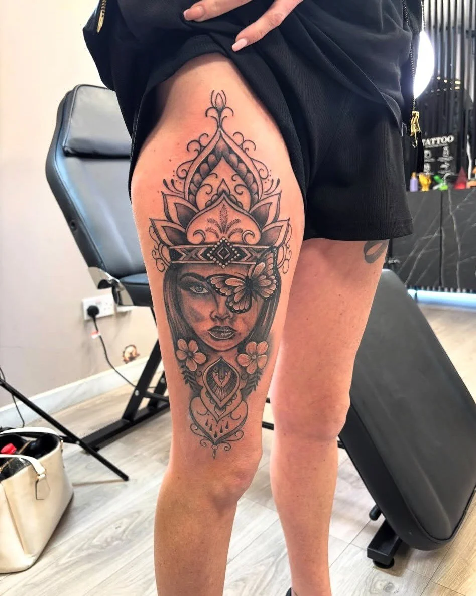 Black and gray tattoo of a woman's face with a butterfly over one eye, surrounded by intricate ornamental designs and floral elements on a person's thigh.