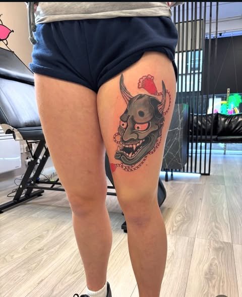 Tattoo of a fierce demon mask on the upper thigh of a person wearing dark shorts.