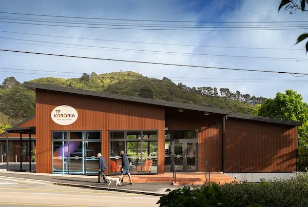 A modern, wooden building with large glass windows, sign reading 'Te Kororia,' situated near a hillside with greenery and power lines in the background.