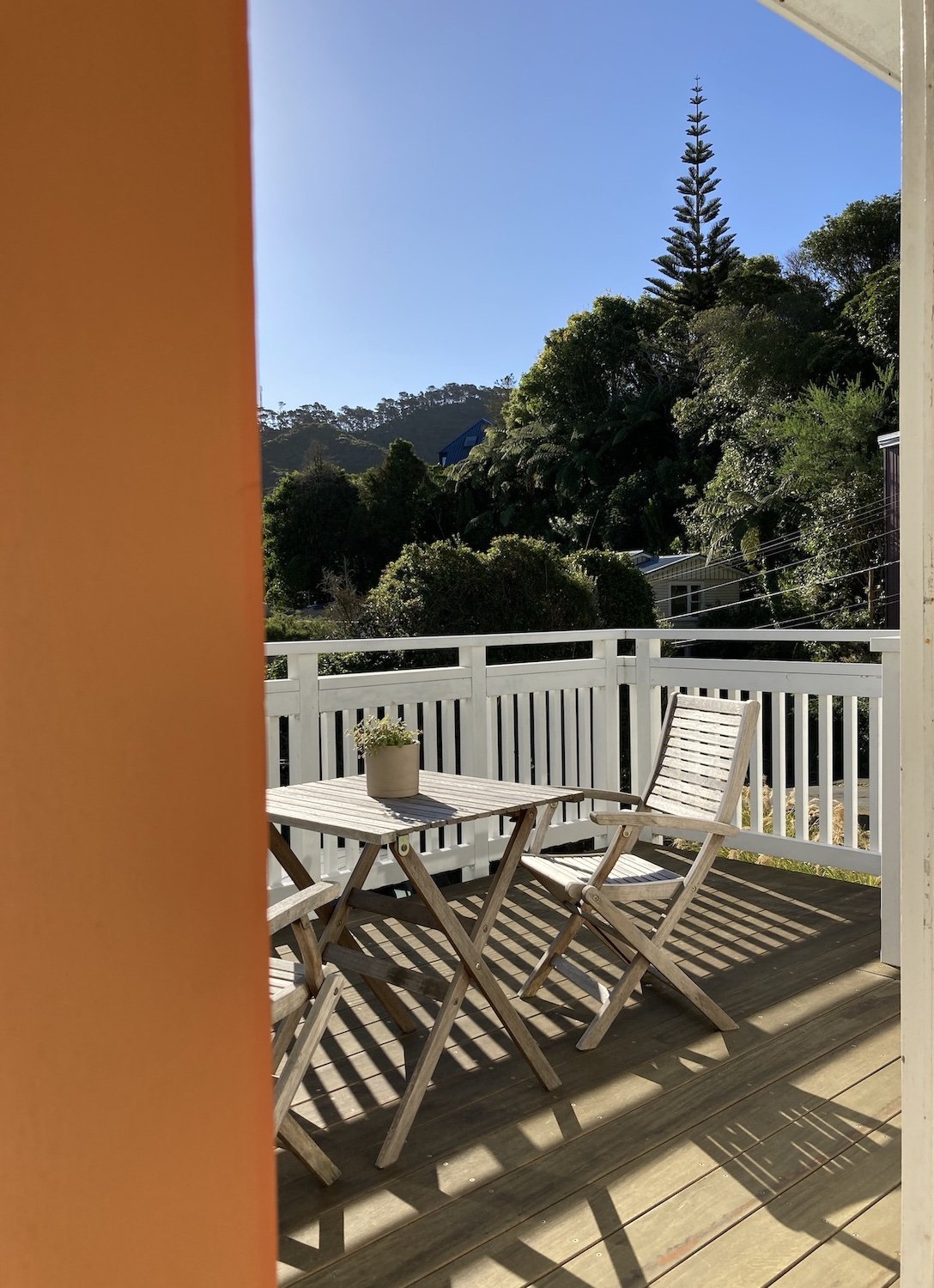 A view of a balcony with white wooden railing, a small wooden table, a wooden chair, and a potted plant, overlooking lush green trees and a clear blue sky.