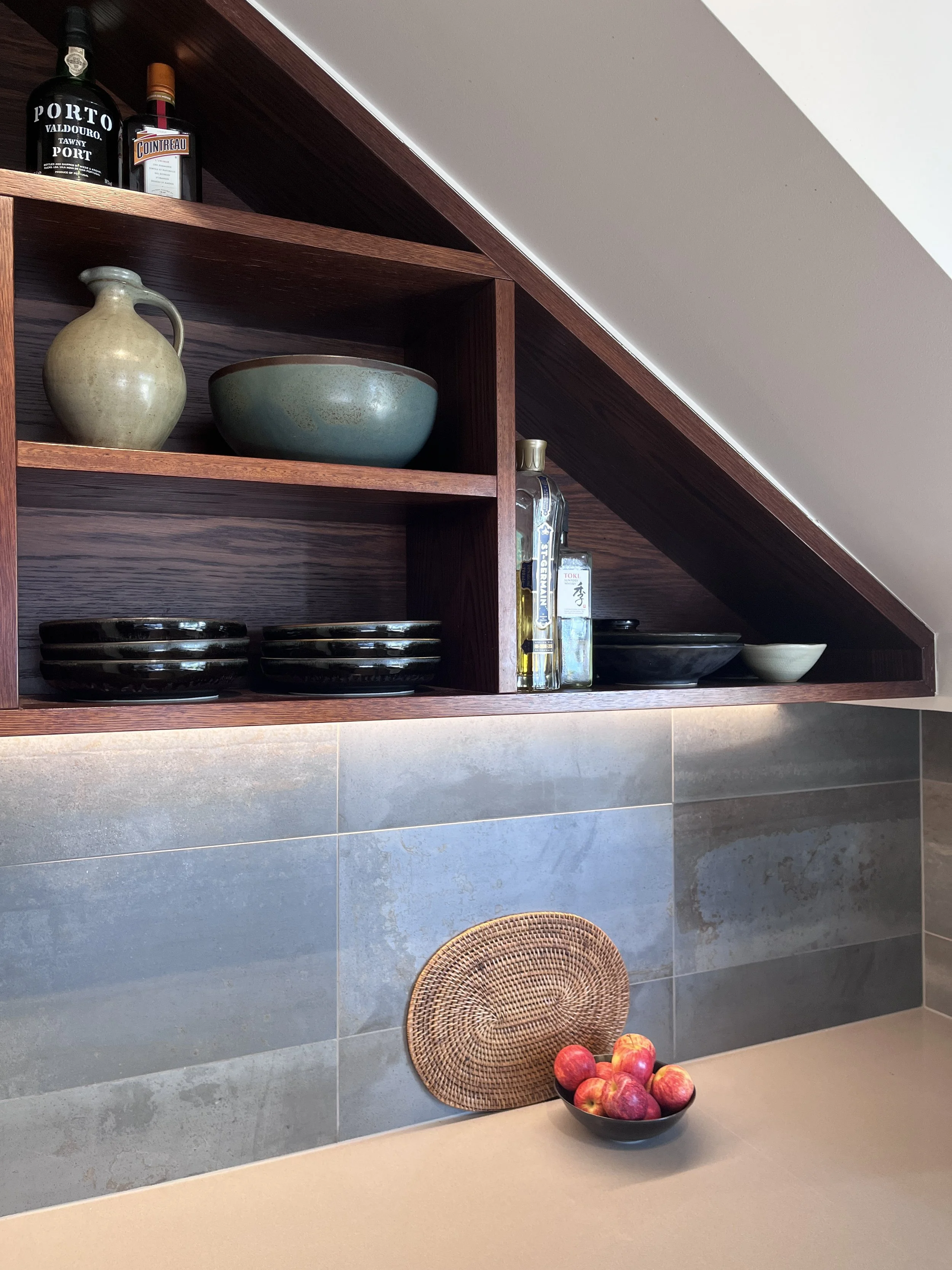 Kitchen countertop with a black bowl of red apples, a woven placemat, and a tiled wall with shelves holding bottles, bowls, and decorative items.
