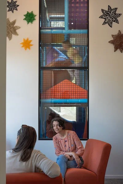 Two women sitting and talking in a room with a large window behind them, decorated with colorful paper snowflakes on the wall.