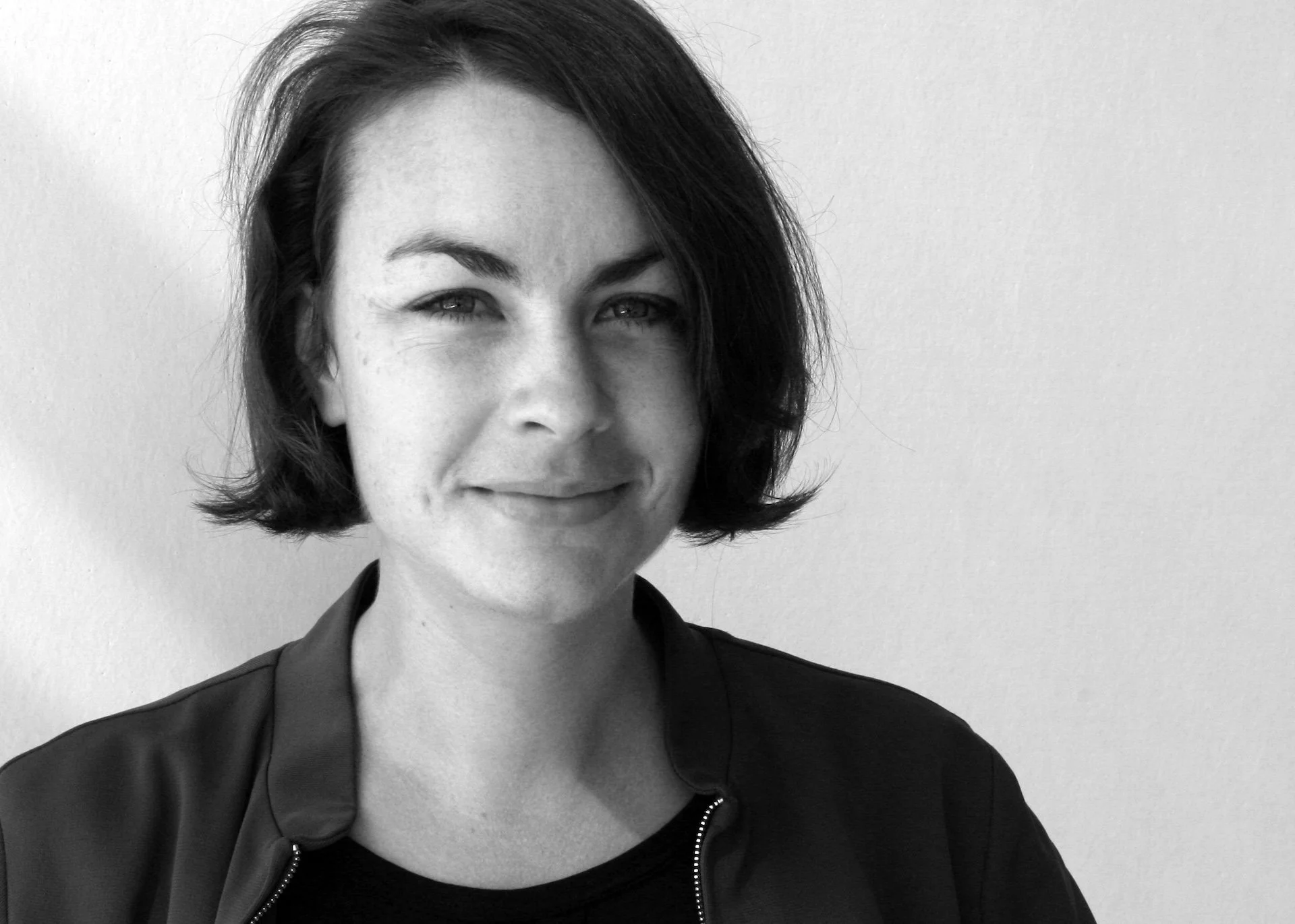 Black-and-white portrait of a woman with short dark hair smiling slightly, wearing a dark top and jacket, against a plain light background.