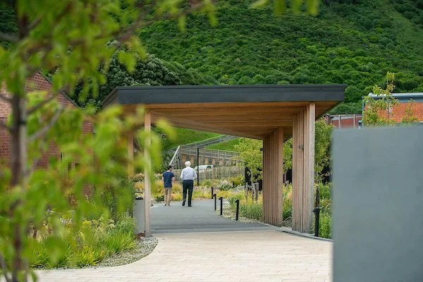 People walking under a modern wooden and metal shelter in a park with lush green hillside in the background.