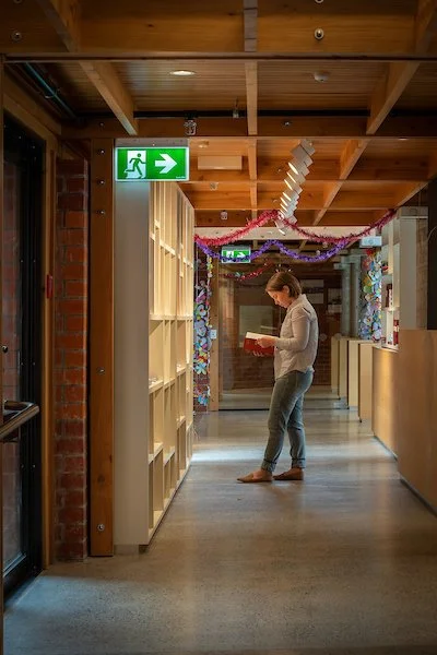 A woman standing in a hallway reading a book or a notebook, decorated with pink and purple hanging streamers, in a building with wood ceiling beams and brick wall, and an illuminated green exit sign with a running figure and an arrow pointing to the right.
