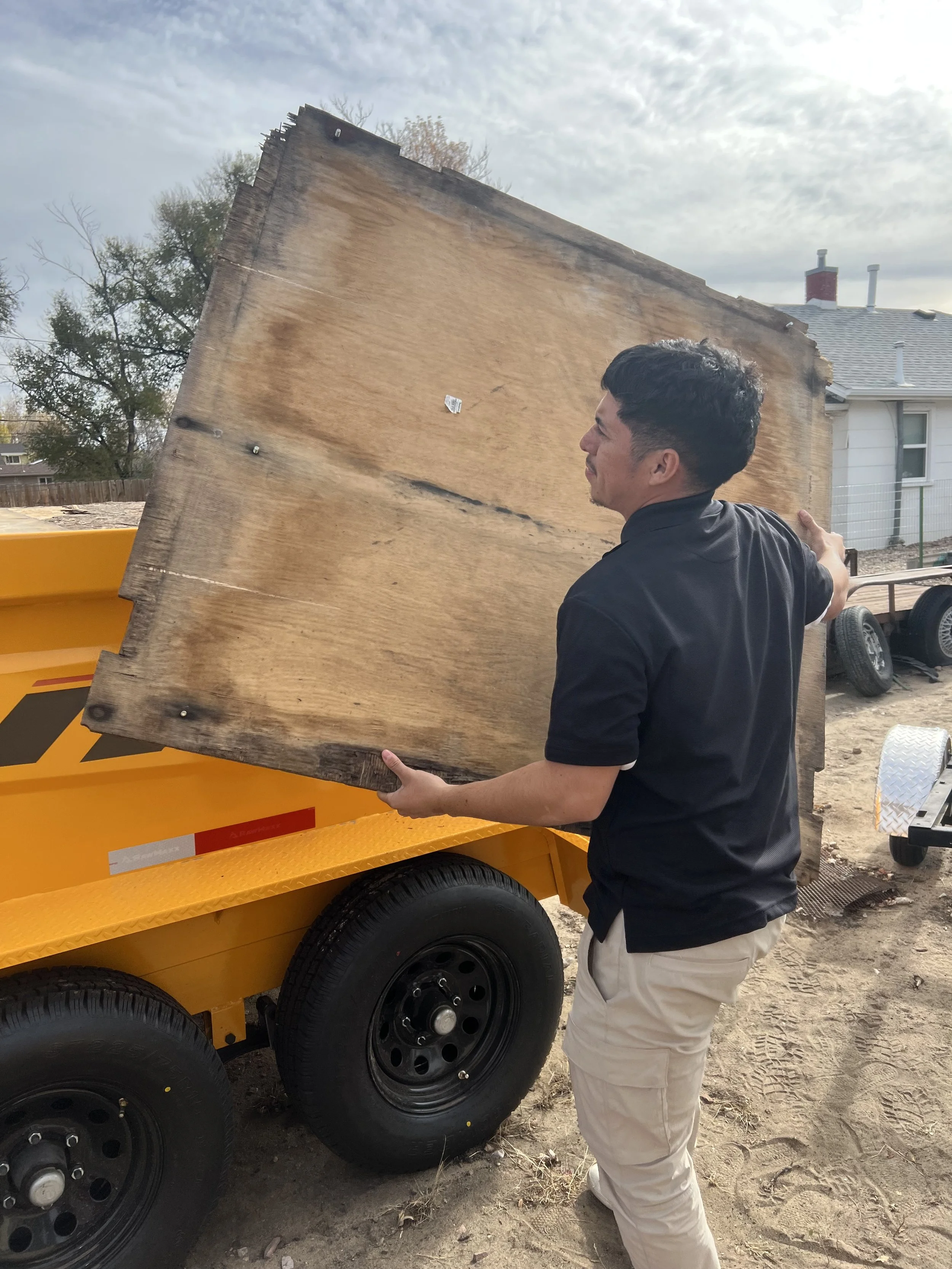 A man in a black shirt placing a large wooden board onto a yellow trailer outdoors.