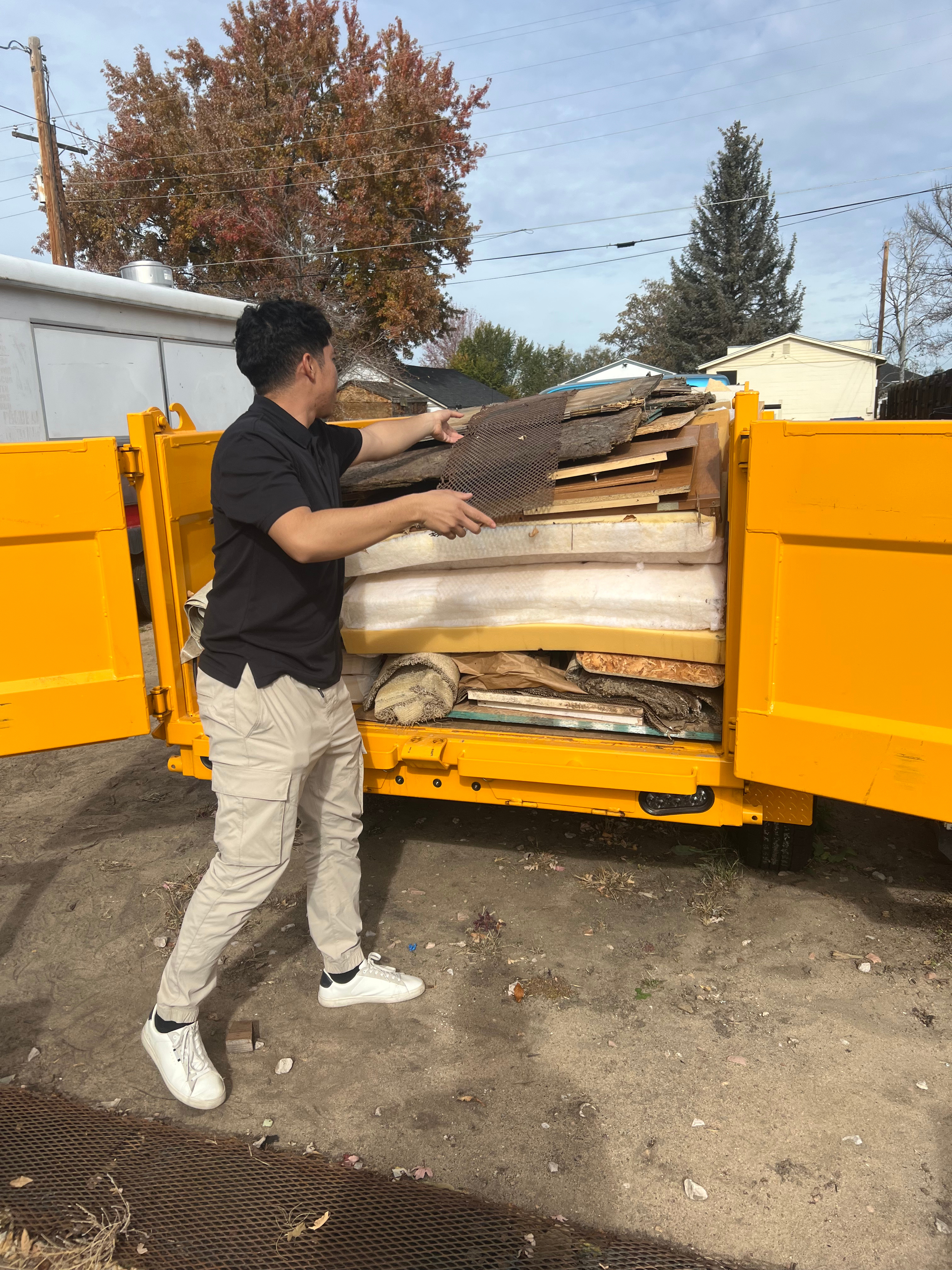 A man in a black shirt and beige pants loading wood and insulation into a yellow truck in an outdoor setting with trees and houses in the background.