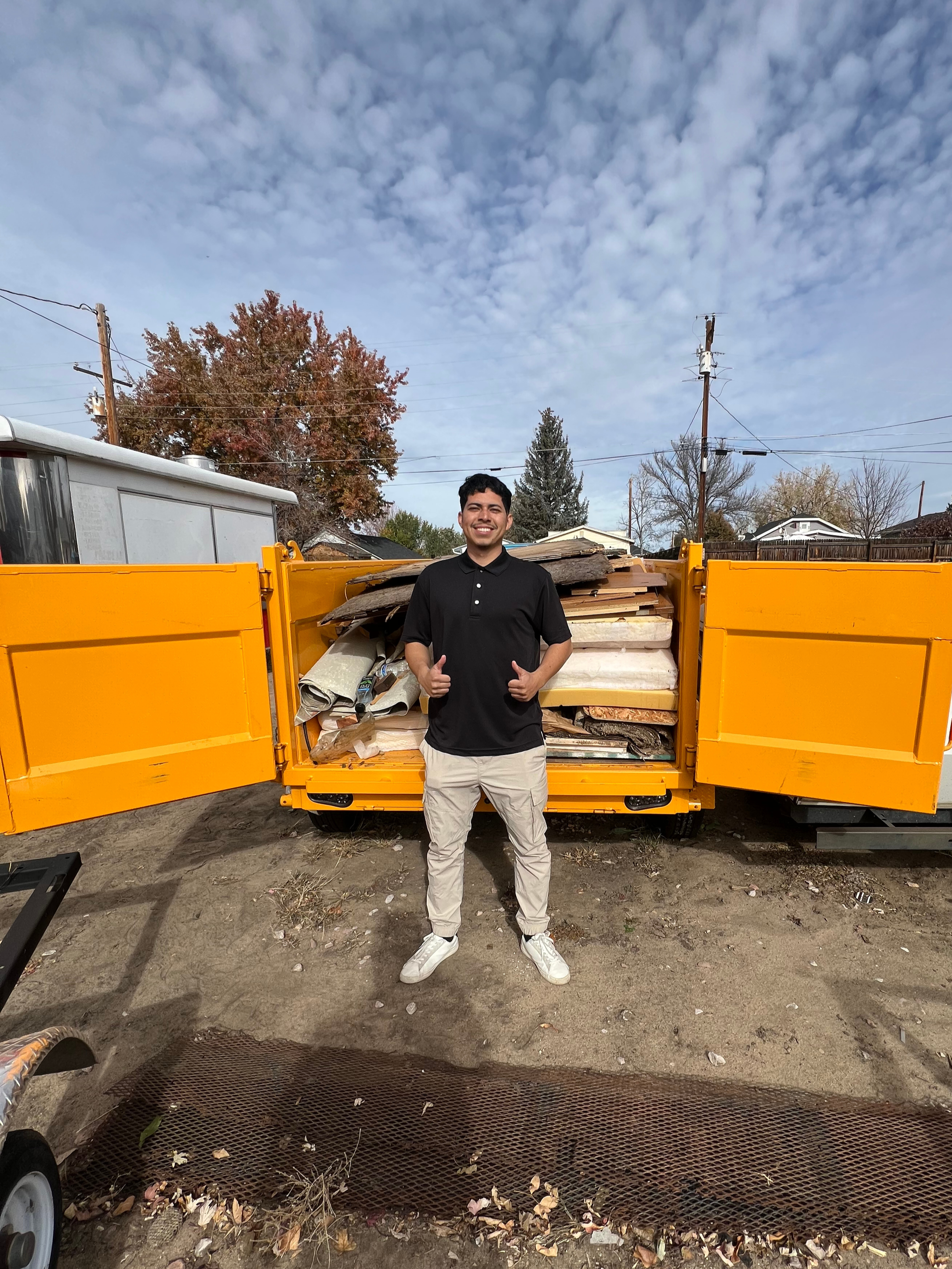 Young man standing in front of a bright yellow dumpster filled with assorted debris, giving a thumbs up and smiling.