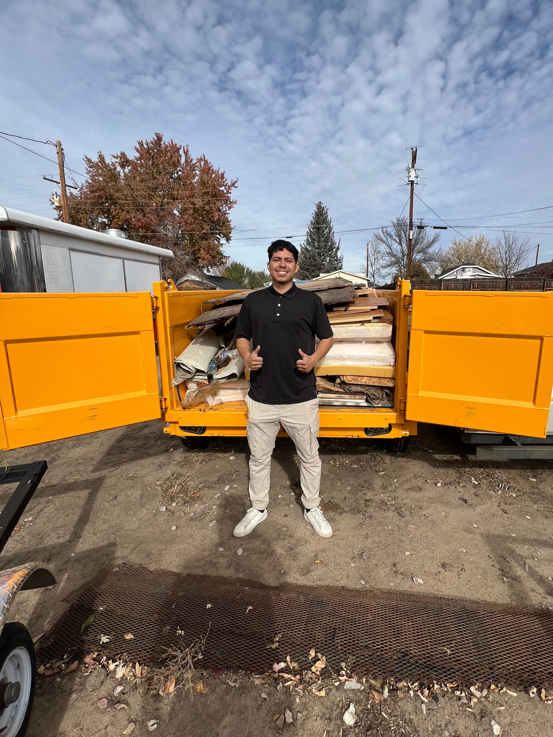 A smiling young man in a black polo shirt and light-colored pants stands in front of an open yellow dumpster filled with debris and wood planks, outdoors on a dirt surface with a cloudy sky overhead.