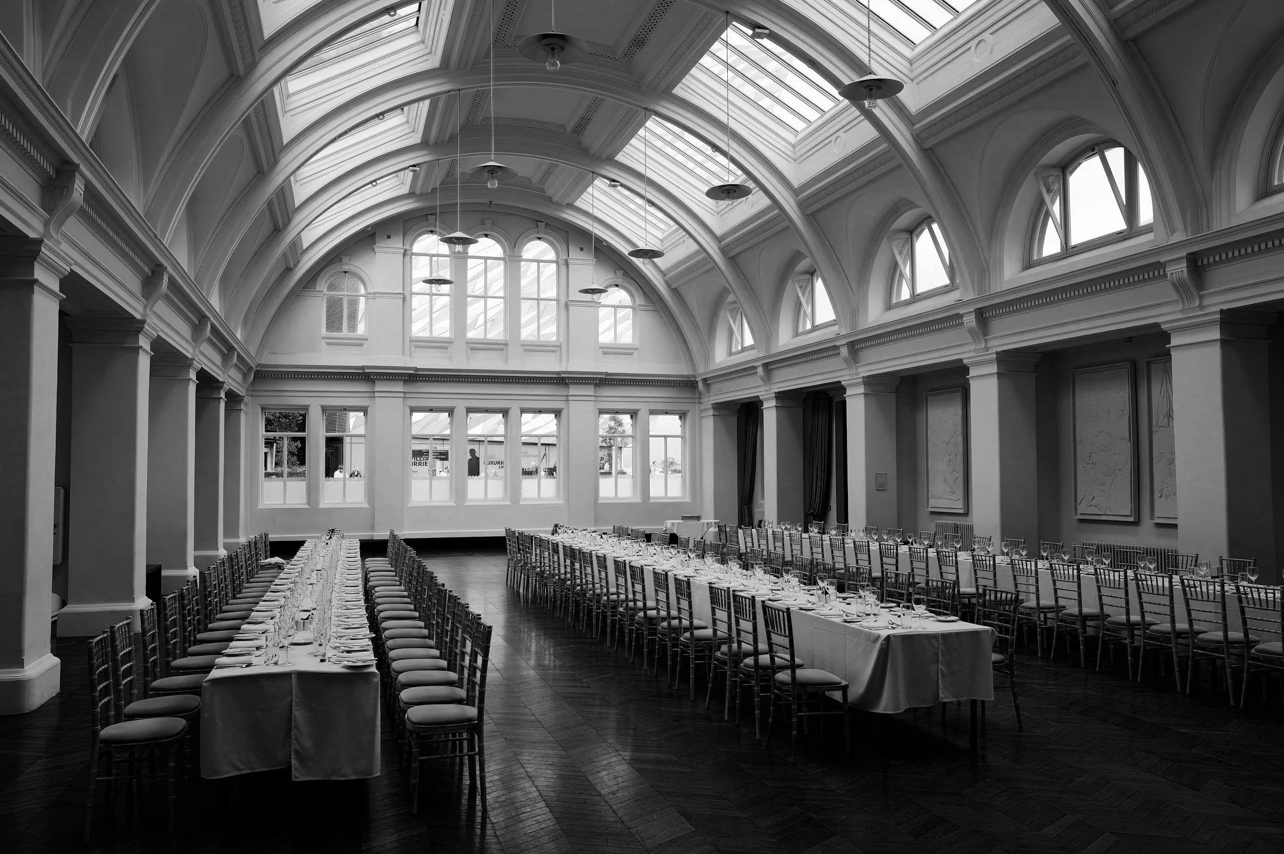 A large banquet hall with high arched ceilings, skylights, and a long, elegantly set dining table with multiple chairs, glassware, and silverware. The room has decorative moldings and tall windows.