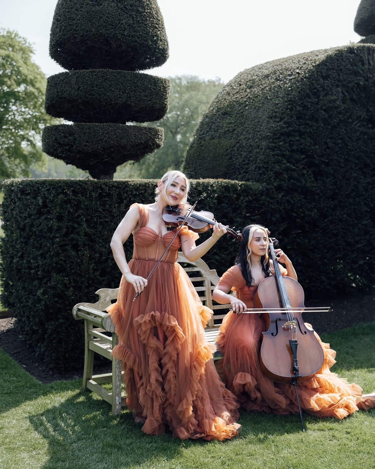 Two women in flowing, ruffled, orange dresses playing classical string instruments in a lush garden. One is standing and playing the violin, while the other is seated and playing the cello. They are outdoors with manicured hedges and a large, sculpted shrub in the background.
