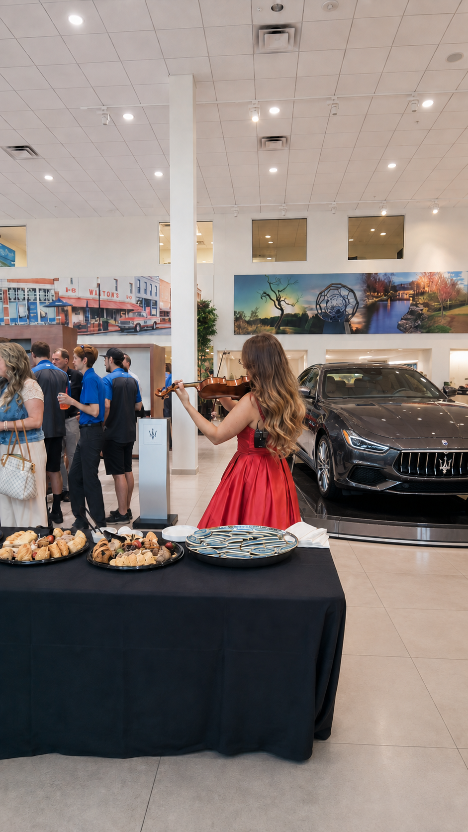 A woman in a red dress playing the violin at a car showroom event with a black Maserati and a group of people in the background.