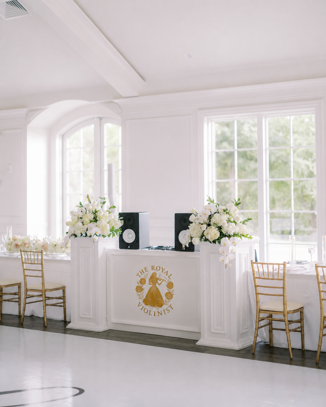 Elegant indoor event space decorated with white flowers, gold chairs, and a DJ setup with speakers, featuring a sign that reads 'The Royal Violist'.