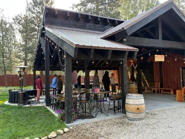 People gathering under a covered outdoor patio with string lights, wooden furniture, and a wine barrel, at a rustic venue with trees in the background.