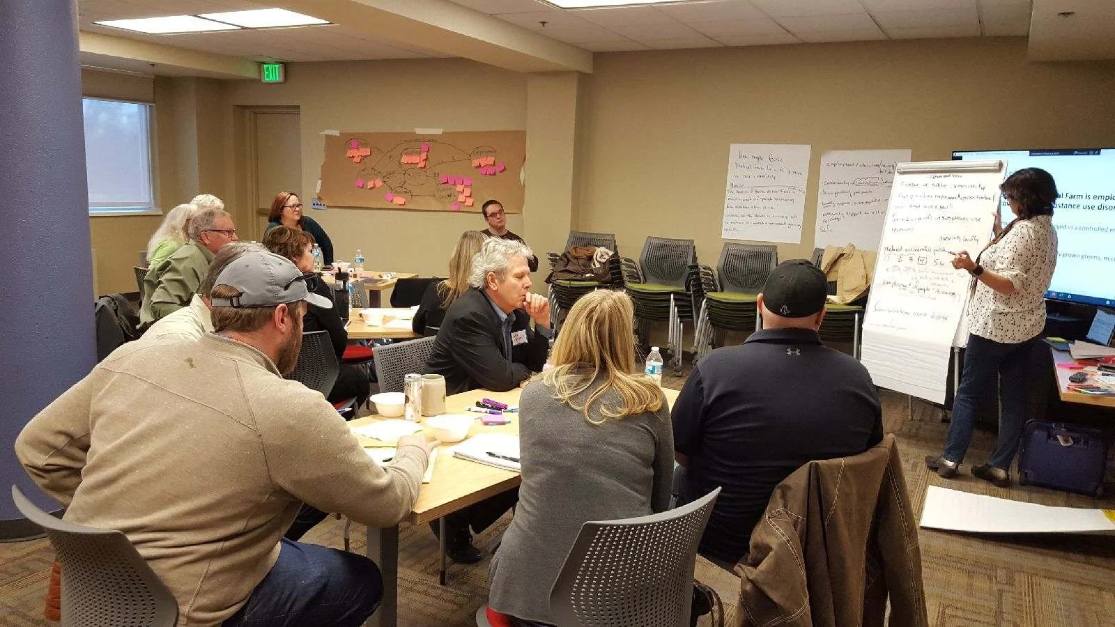 Group of people in a meeting or workshop room, sitting at tables, listening to a woman presenting with notes on a large notepad and a presentation slide on a screen.
