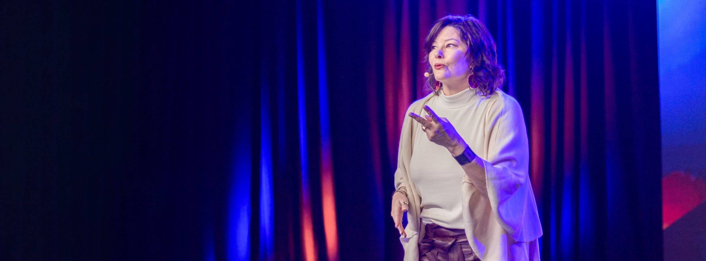 A woman with shoulder-length brown hair and red earrings, wearing a beige top and brown pants, is speaking on stage with a microphone headset, gesturing with her right hand. The background has vertically hanging blue and red curtains illuminated by stage lights.