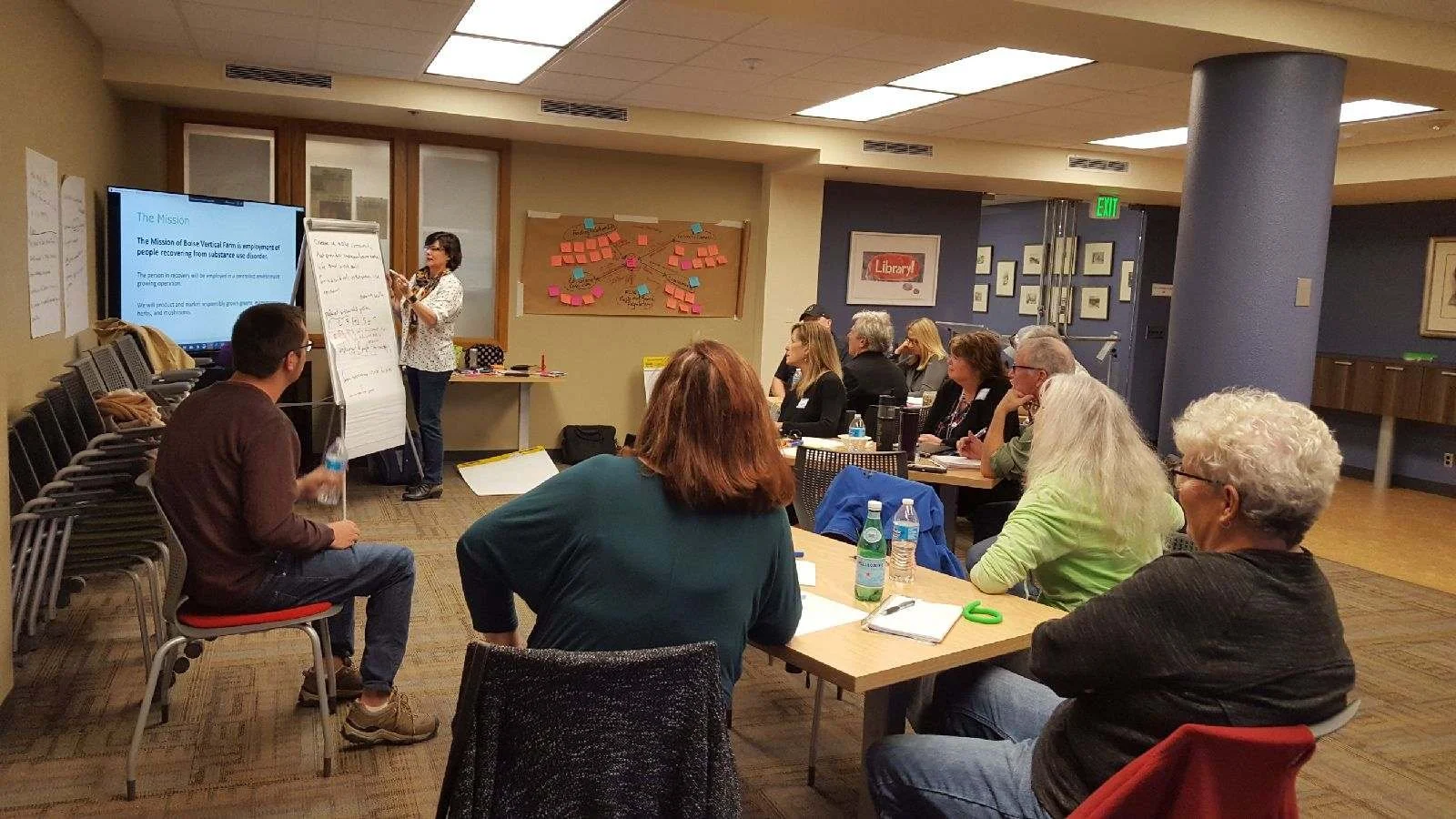 A group of adults attending a business or educational seminar in a conference room, with a woman speaking at a whiteboard and slide presentation.