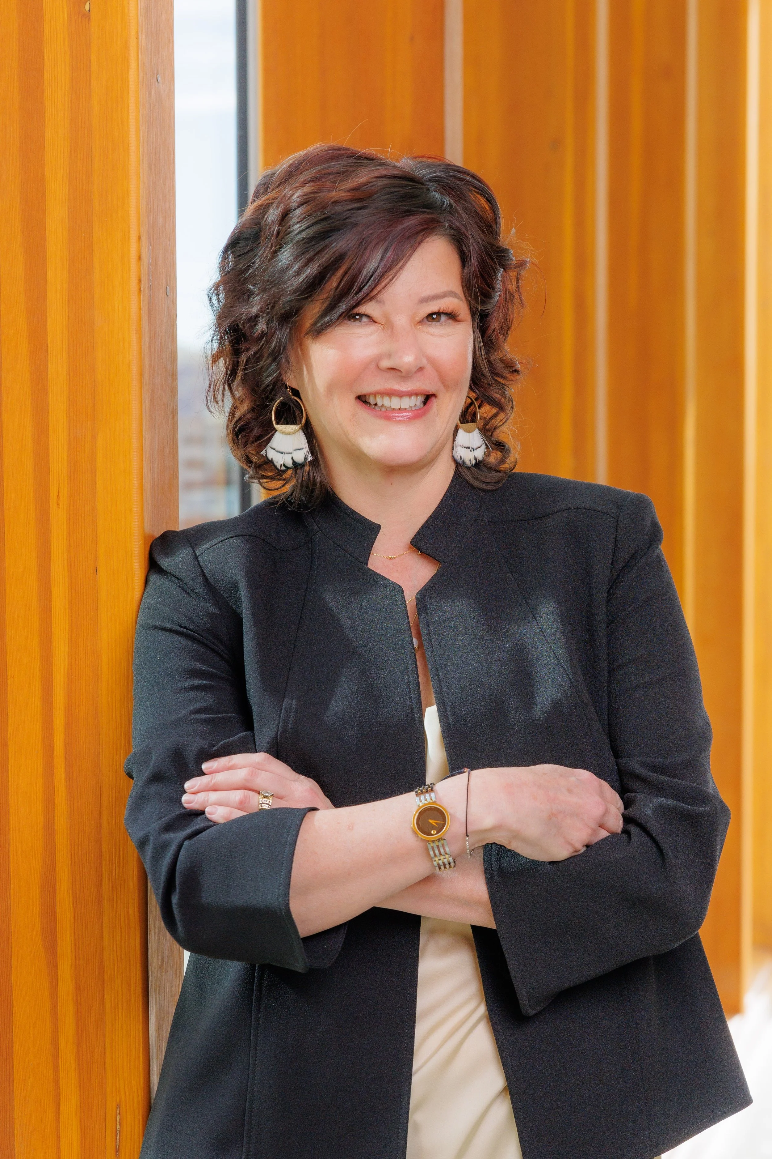 A smiling woman with wavy dark brown hair leaning against a wooden wall, wearing a black blazer, large circular earrings, a wristwatch, and a bracelet.