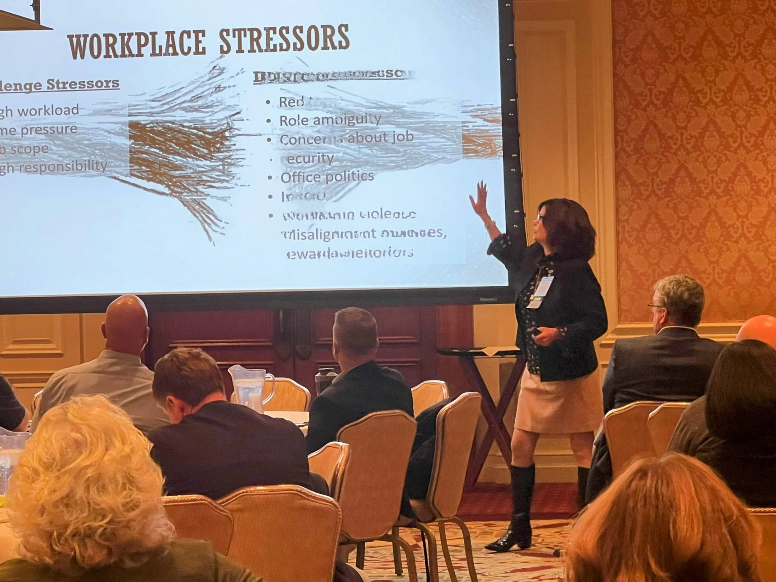 A woman in a black blazer and knee-high boots giving a presentation on workplace stressors to an audience in a conference room with ornate wallpaper. The presentation slide behind her shows a tree diagram and a list of stressor types.