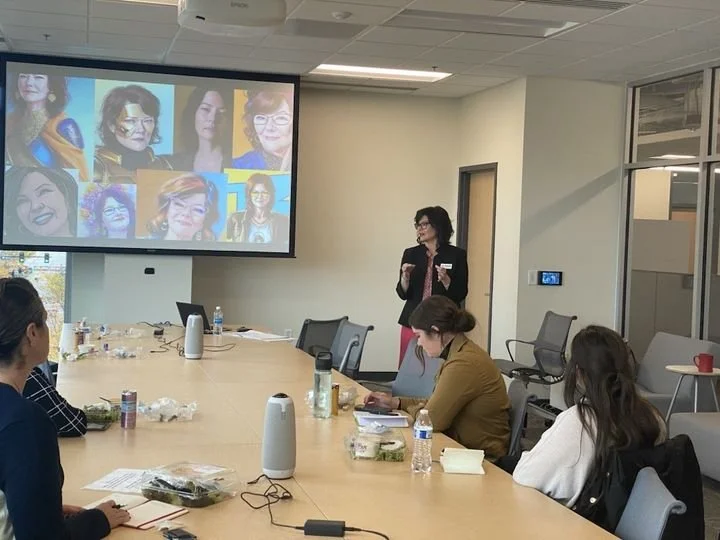 A woman is giving a presentation to a group of people in a conference room. The presentation slide shows multiple portraits of women. Attendees are sitting around a large table with water bottles, notebooks, and snacks.