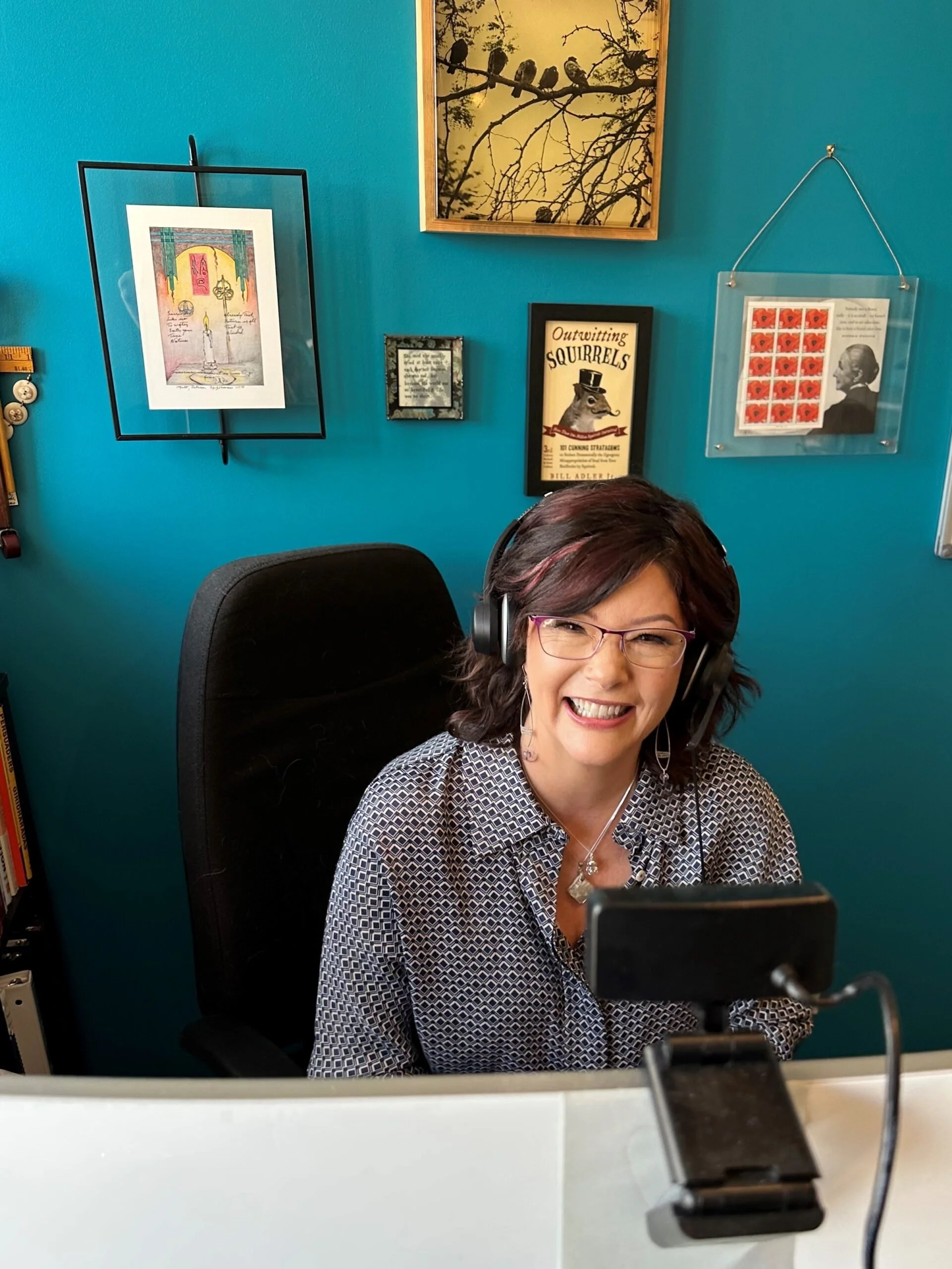 A woman wearing glasses and headphones sitting at a desk, smiling and looking at a smartphone mounted on a stand in front of her. Behind her is a blue wall decorated with framed artwork and posters.