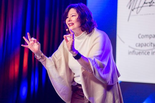 Woman speaking on stage with a presentation slide in the background, gesturing with her hands.