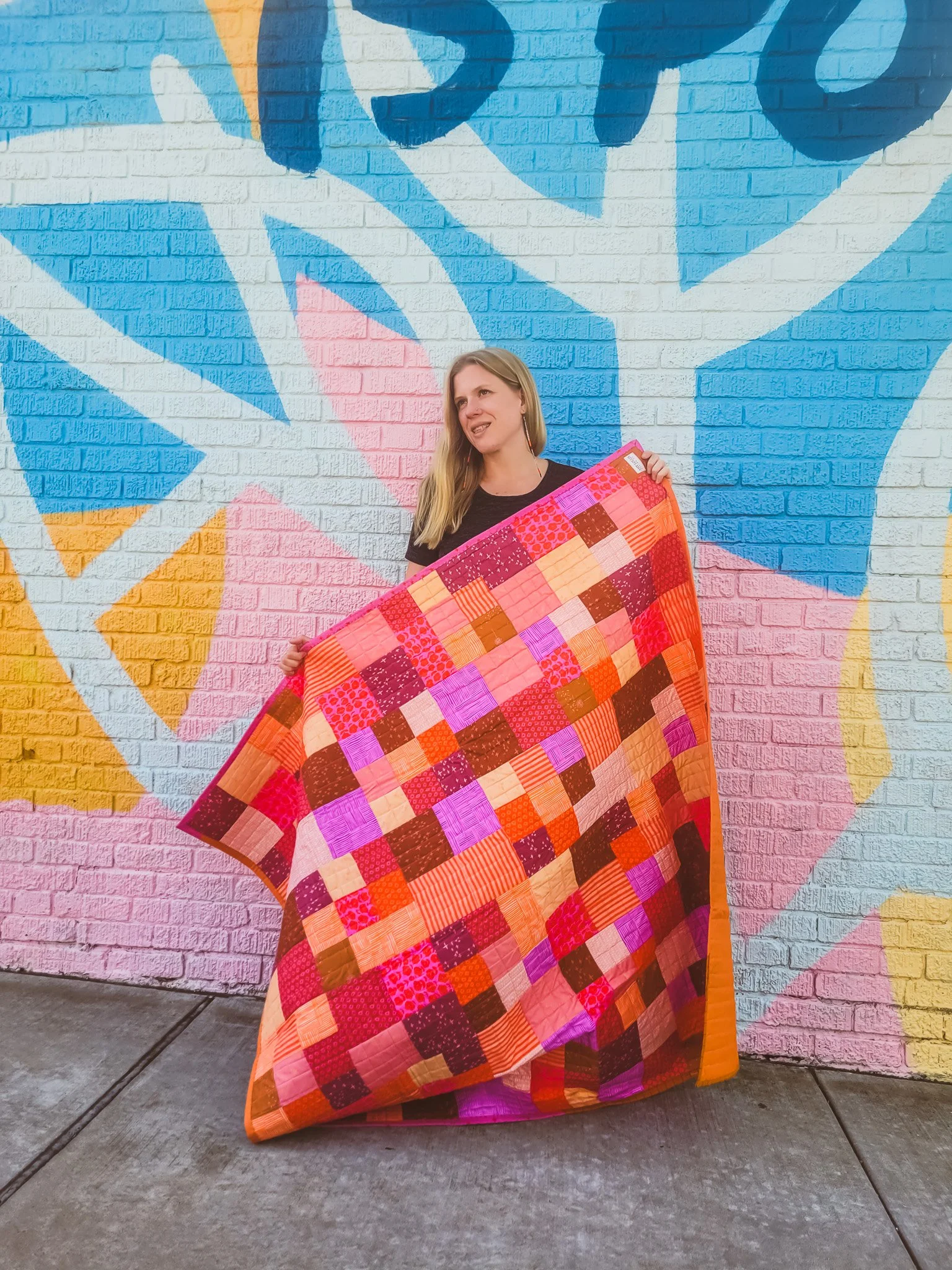 A woman standing in front of a colorful graffiti mural, holding a vibrant patchwork quilt with pink, red, and orange squares.