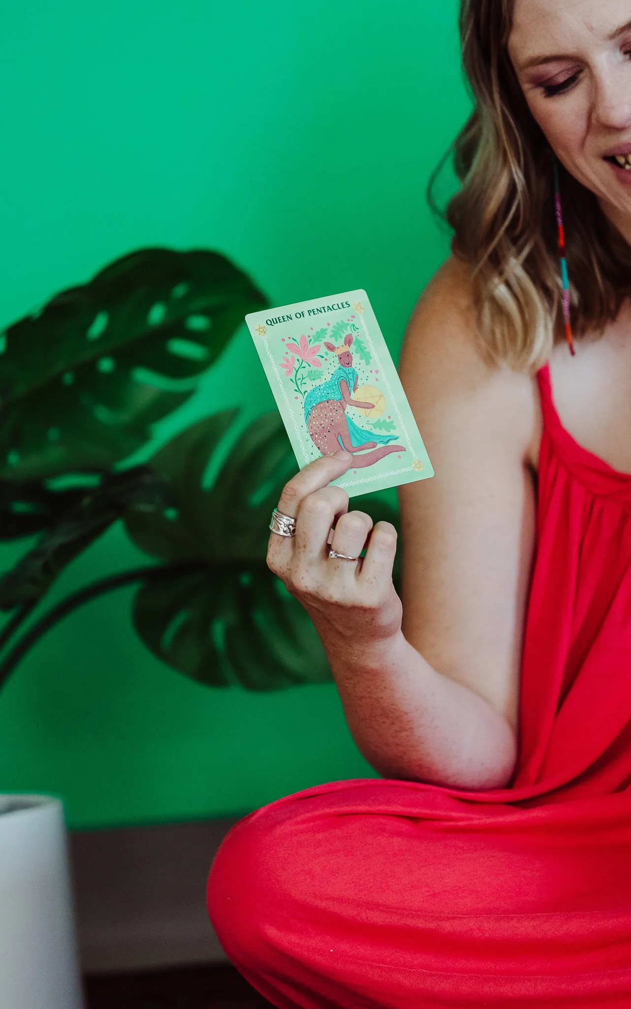 A woman in a red dress is holding up a tarot card titled Queen of Pentacles, with a colorful illustration of a deer with flowers | Amy Wilder Studio - Brand Strategist + Web Design