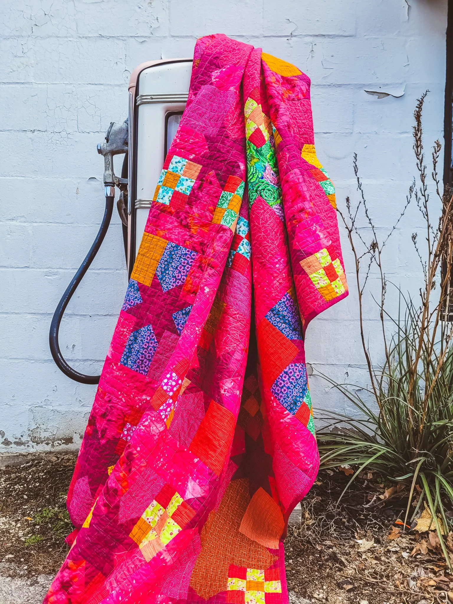 A vibrant pink quilt with various colorful patchwork patterns draped over a vintage white refrigerator outdoors, against a textured white wall with some dried plants on the side.