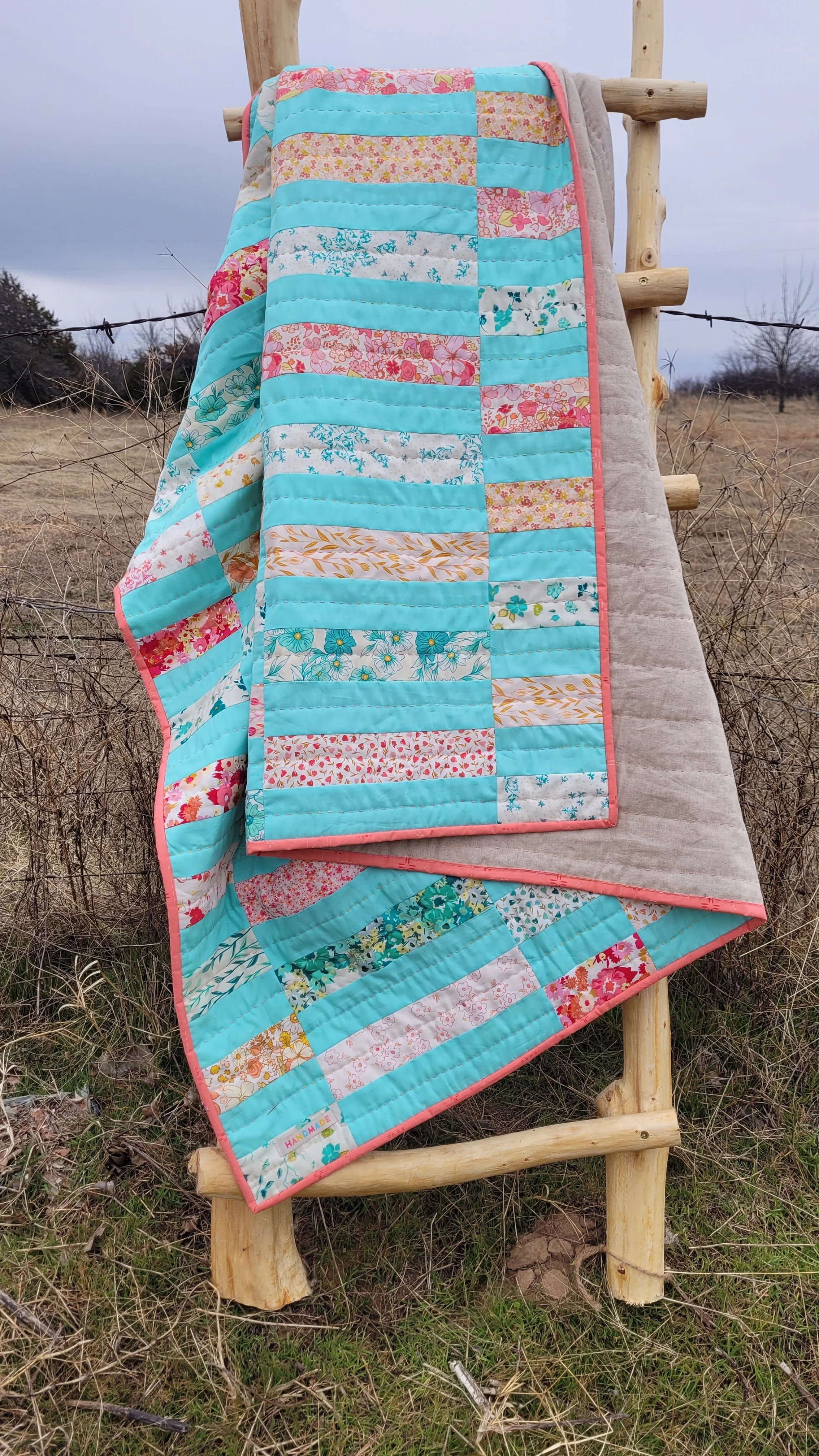 A colorful patchwork quilt with various floral and solid fabric patterns, draped over a wooden ladder outside in a field with dry grass and trees in the background.