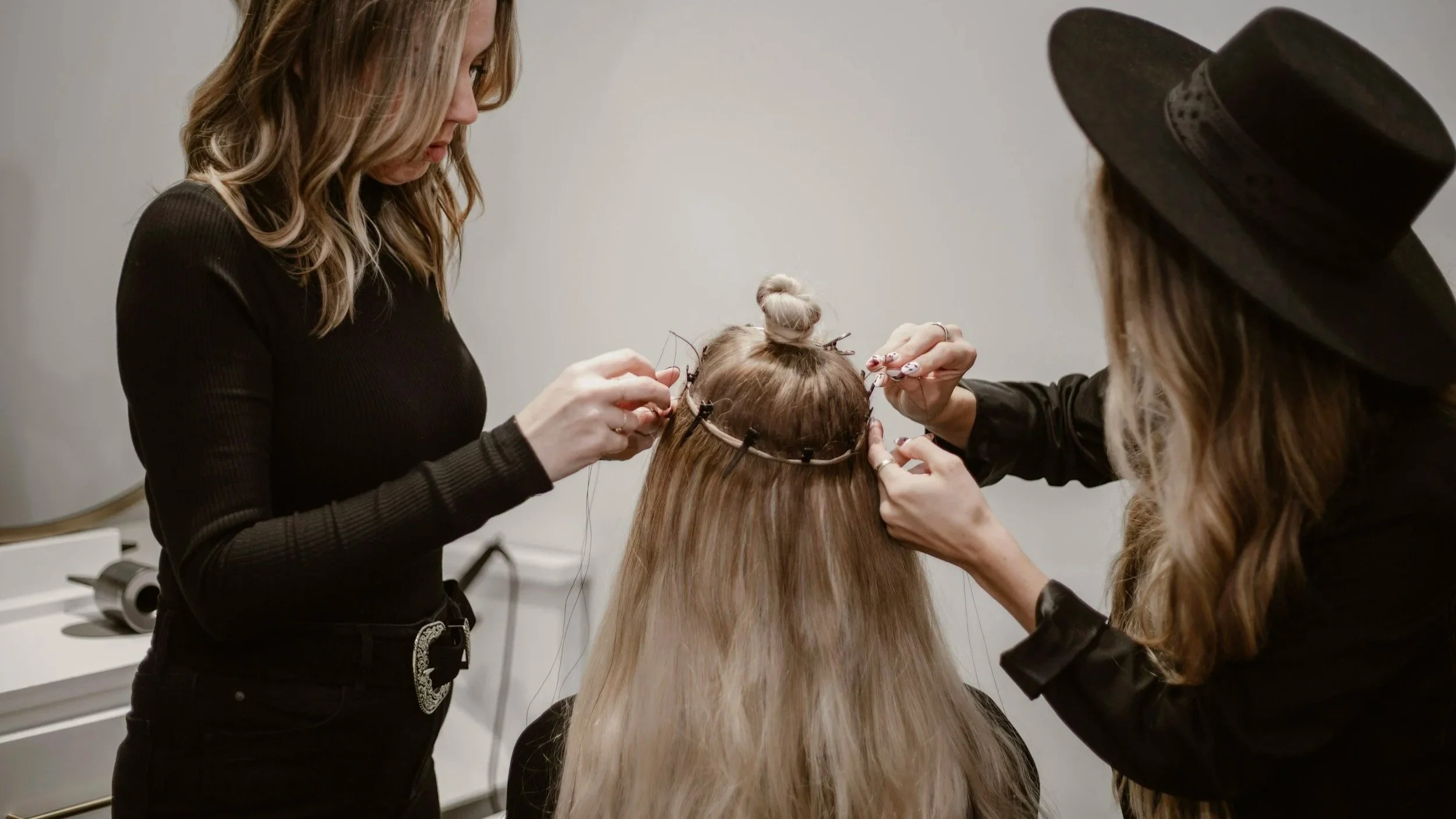 Two women are working on a woman's hair, making a hair bun on top of her head, while she wears a decorative headband, in a salon setting.