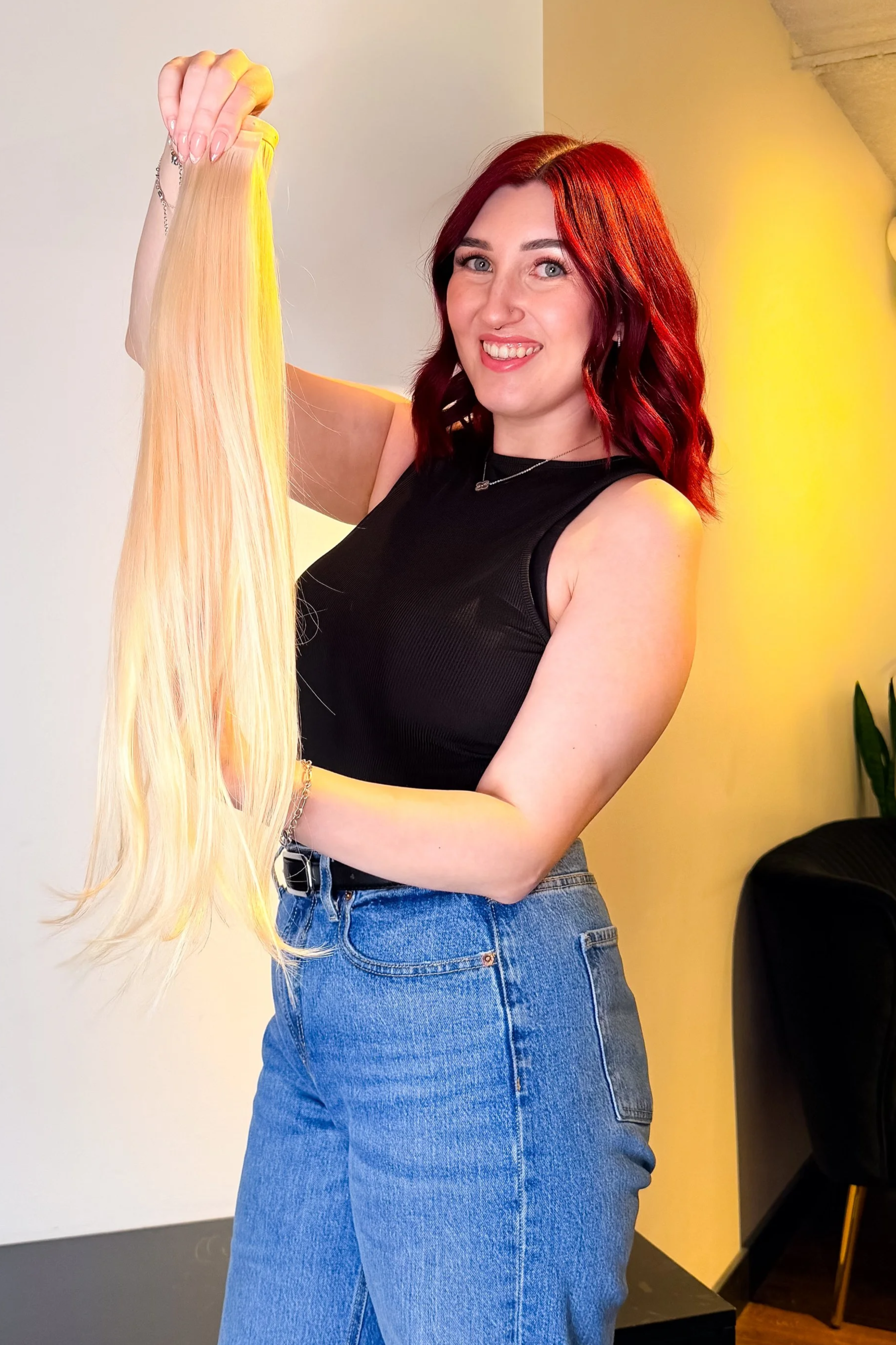 Woman with red hair holding a blonde hair extension in a brightly lit room.