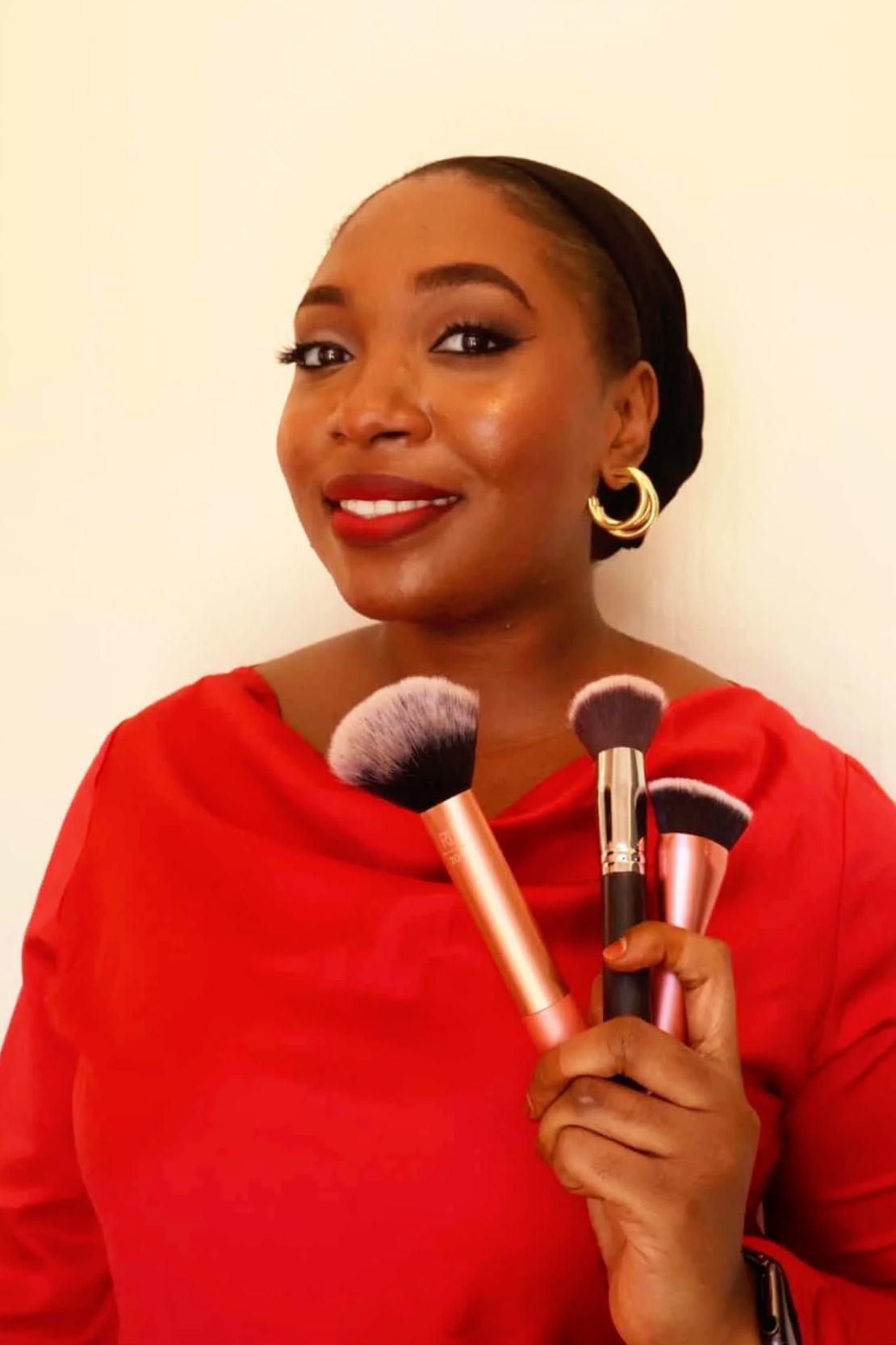 A woman with makeup, gold earrings, and a light purple headscarf, wearing a tank top and a gold necklace, standing in front of window blinds.