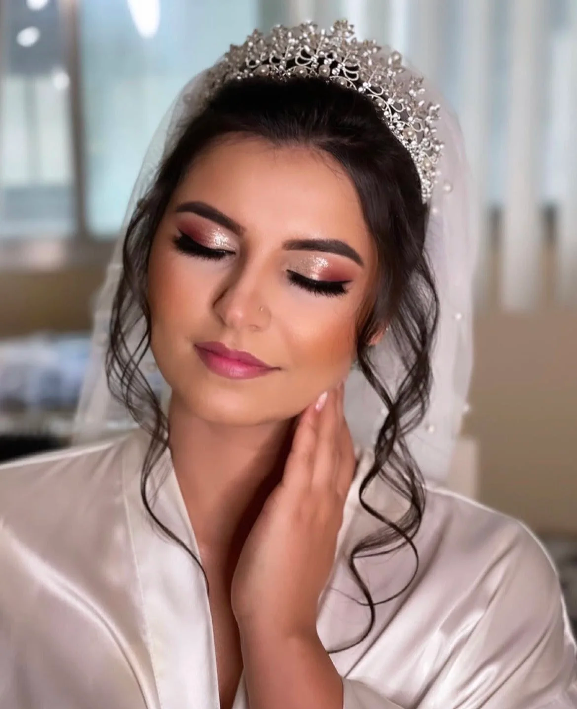 A bride with closed eyes, wearing a tiara and a satin robe, with makeup and a serene expression.
