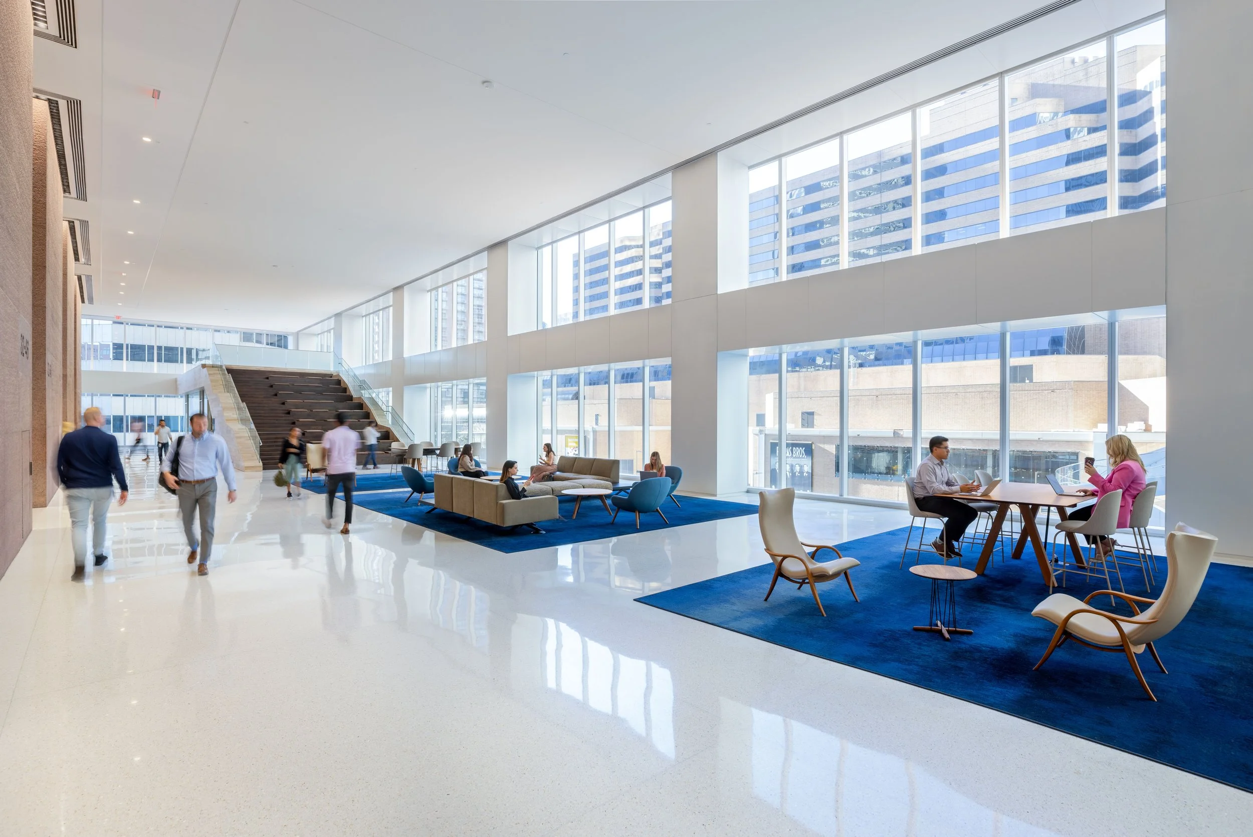 Modern lobby with large windows, blue rugs, seating area with couches and chairs, and people working or walking around.