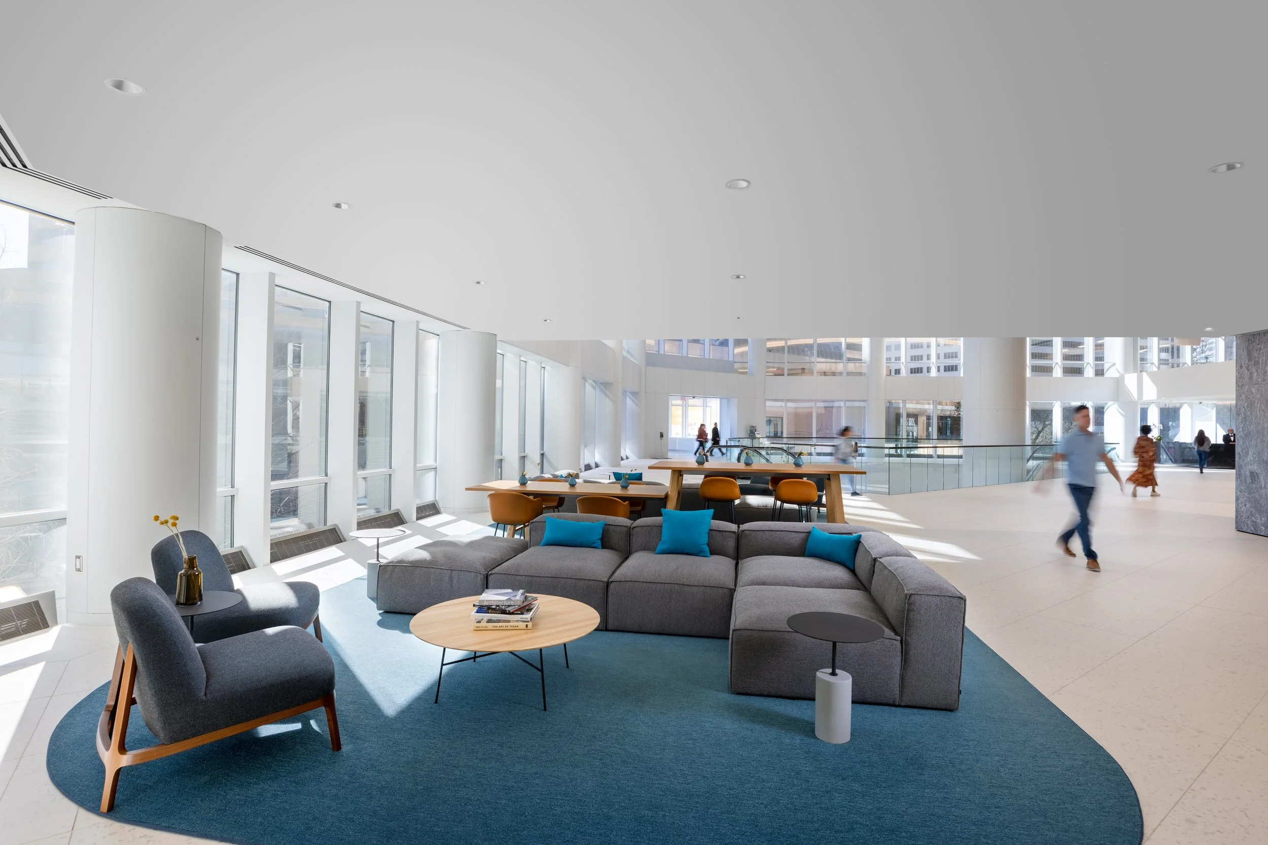 Modern, bright lobby with large windows, gray seating area with blue cushions, a round wooden coffee table with magazines, two gray armchairs with wooden legs, and a blue rug. In the background, there are high tables with orange chairs, and people walking through the space.