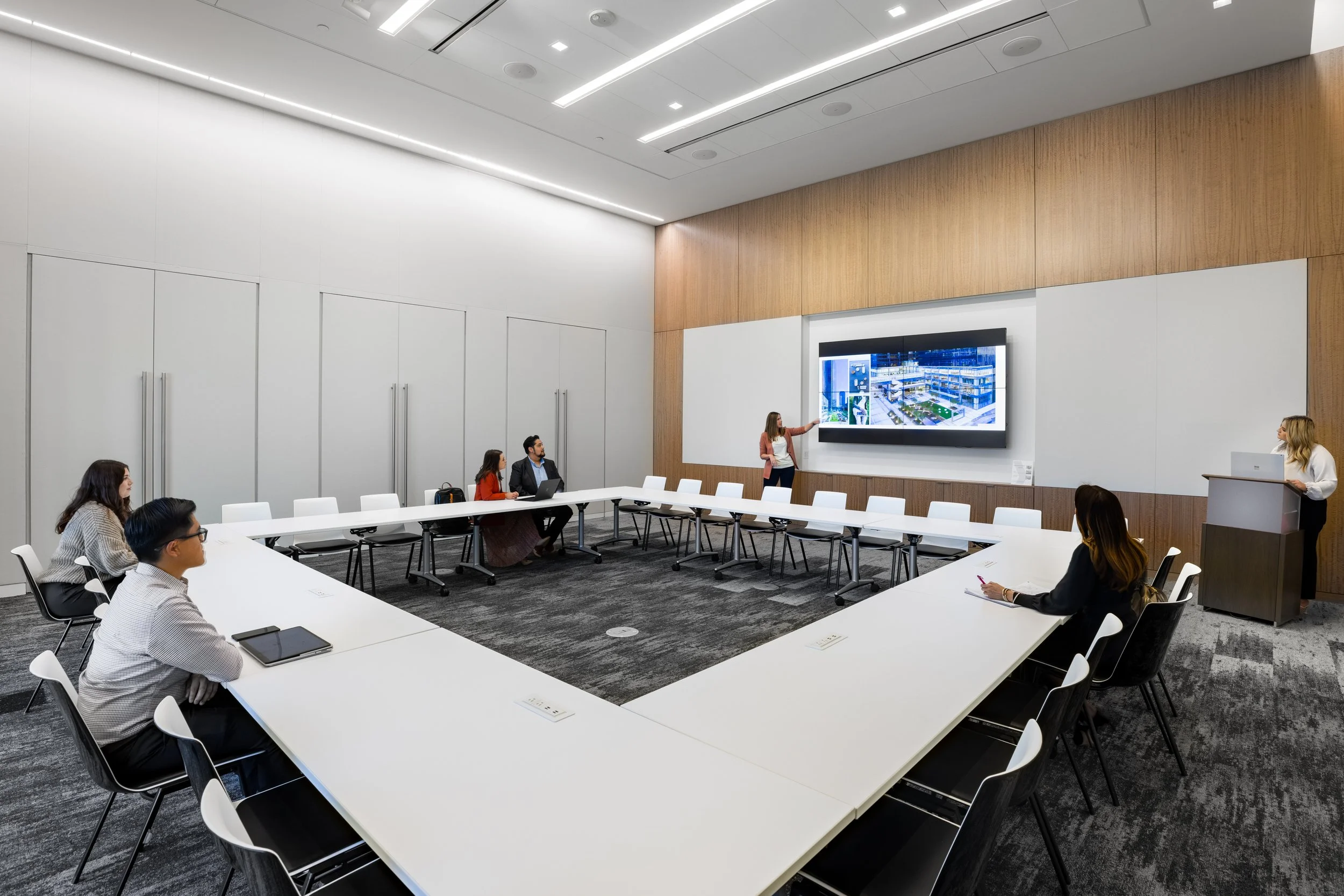 Business meeting room with people seated around a U-shaped conference table, a woman giving a presentation with a large screen on the wall, and a woman at a podium in the corner.