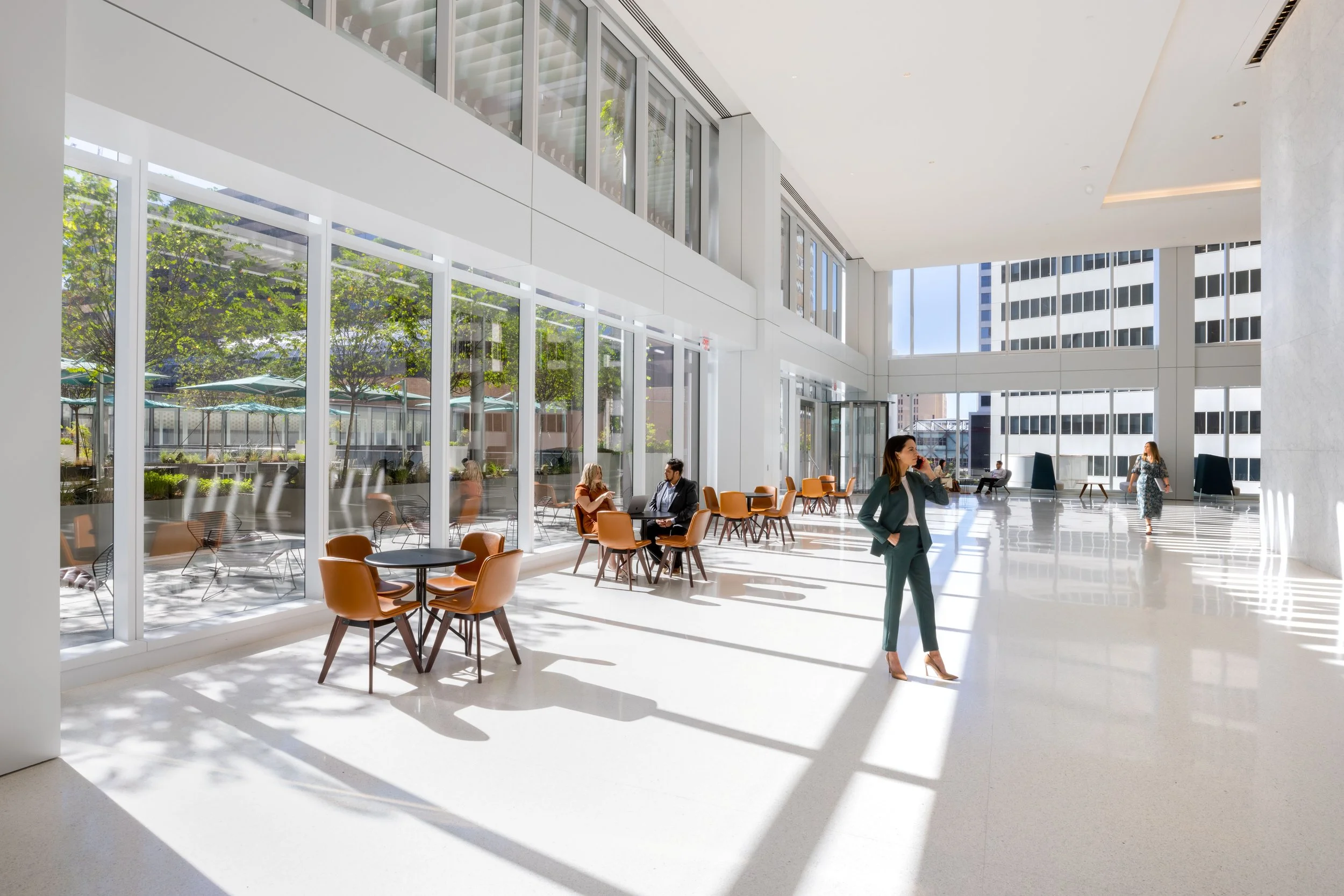 Modern office lobby with floor-to-ceiling glass windows, sunlight streaming in, and several people seated and walking.