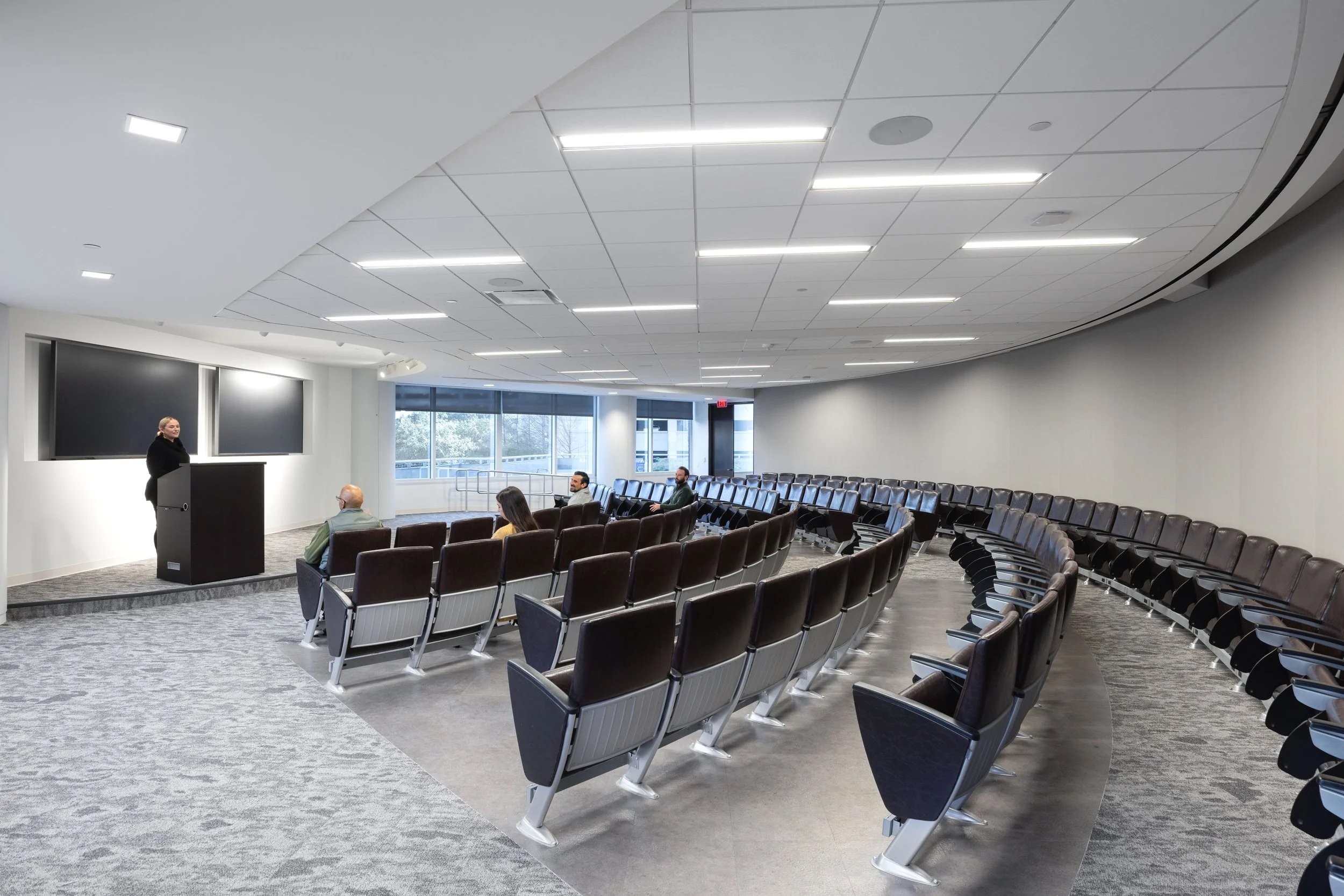 A conference room with a speaker at a podium and four audience members seated, with rows of chairs and large windows in the background.