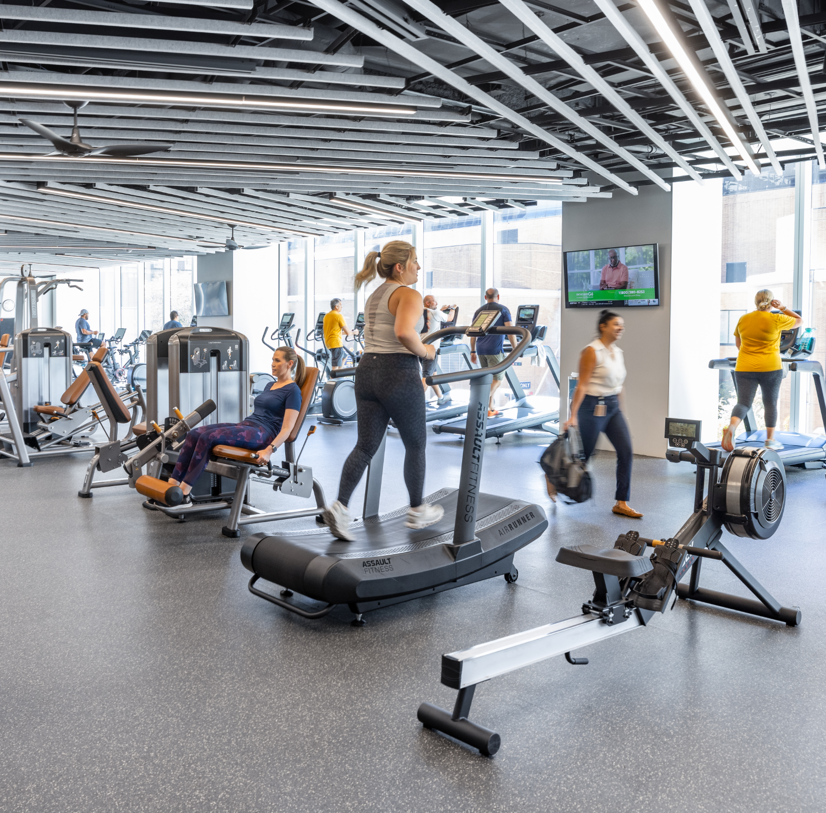 People exercising on cardio machines in a modern gym with large windows, black ceiling with decorative panels, and a TV screen on the wall.