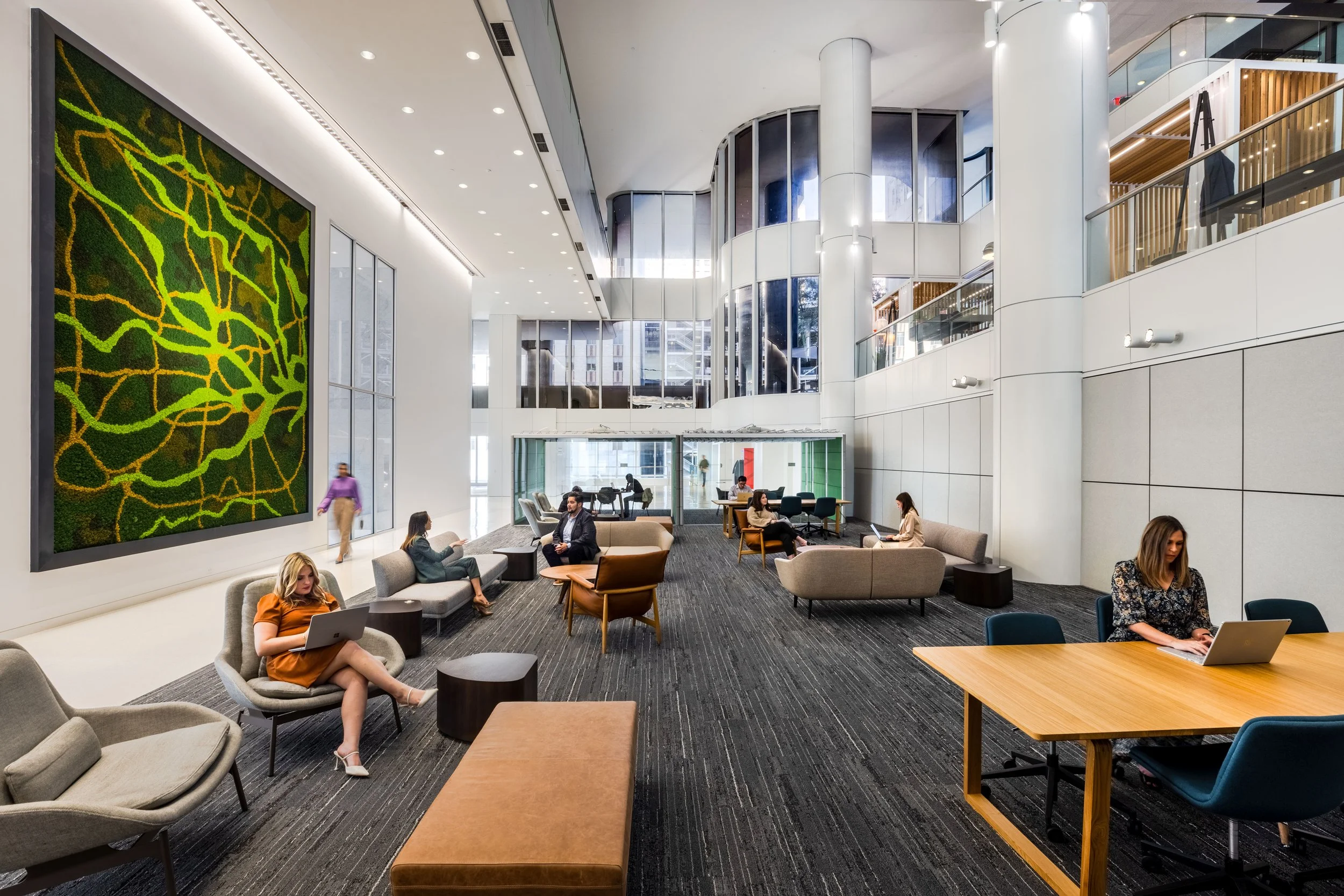 Modern office lobby with people working on laptops, seating areas, large wall art, and glass elevator shaft.