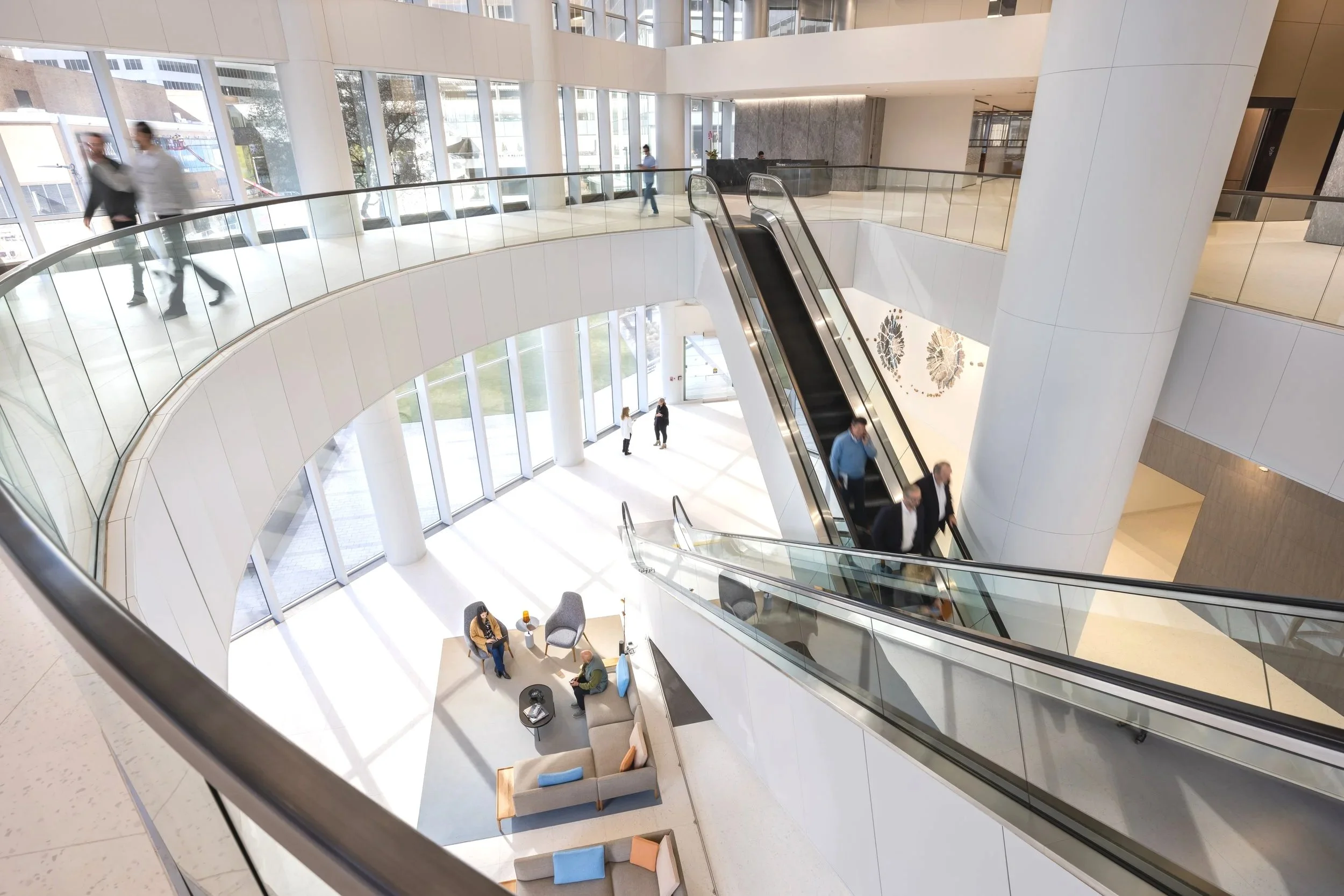 Modern multi-story office building lobby with glass walls, an escalator, and seating area where people are sitting and walking.