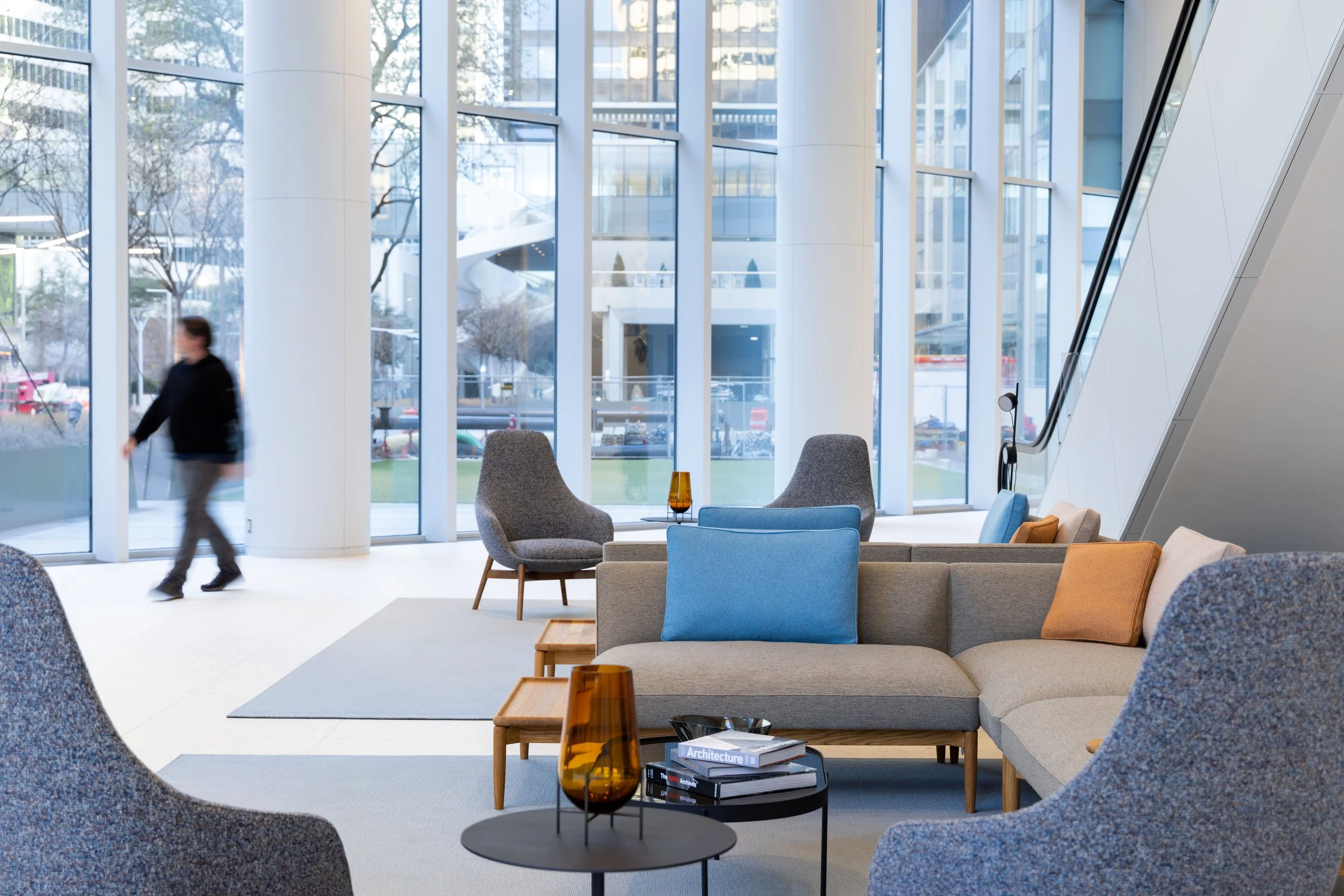 Modern hotel lobby with large glass windows, contemporary furniture including gray armchairs, a beige sofa with colorful pillows, and a black table with books and amber vases. A person is walking outside visible through the windows.