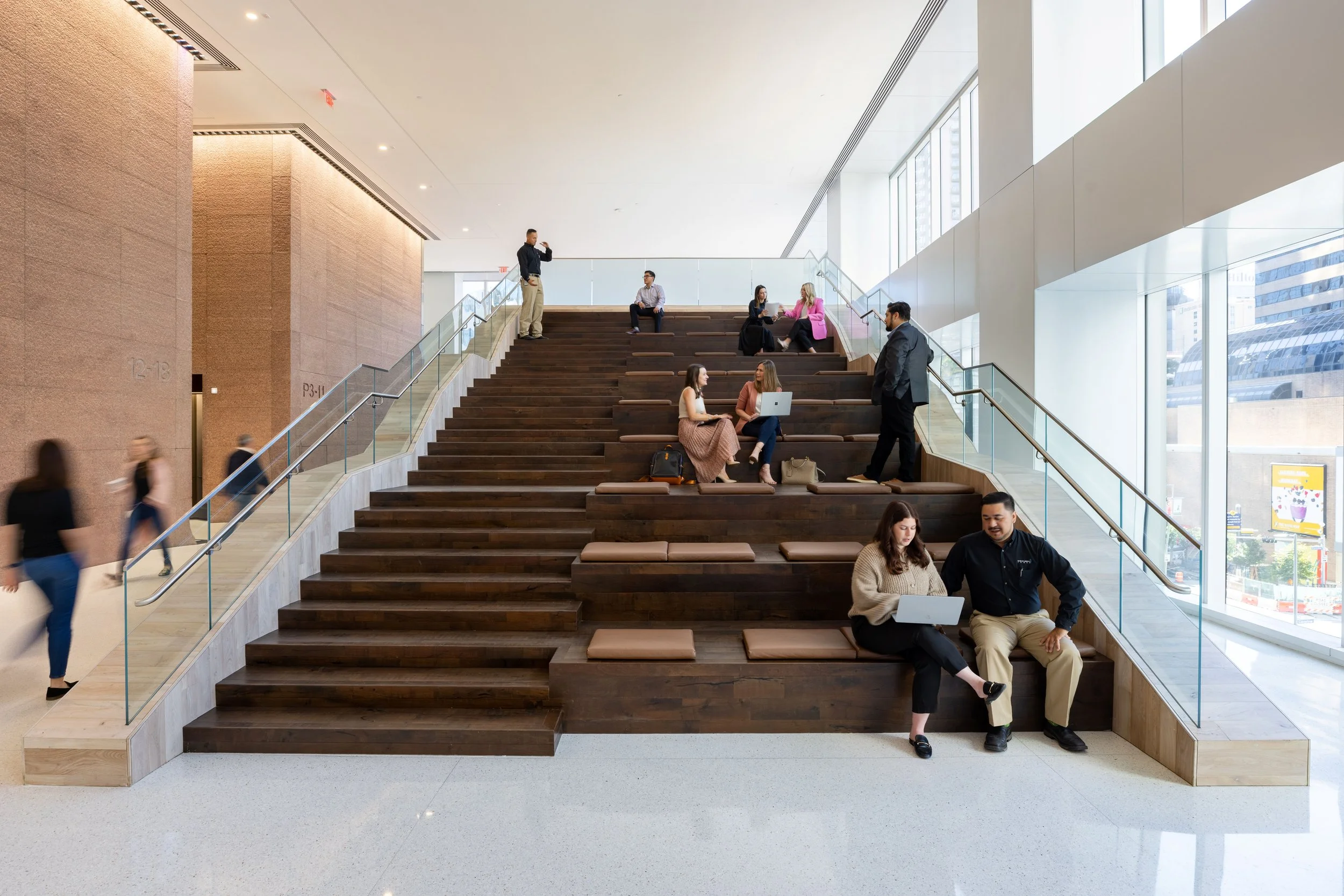 People sitting on a multi-level wooden seating area in a modern building lobby, some working on laptops, others talking, with large windows and an urban street scene outside.