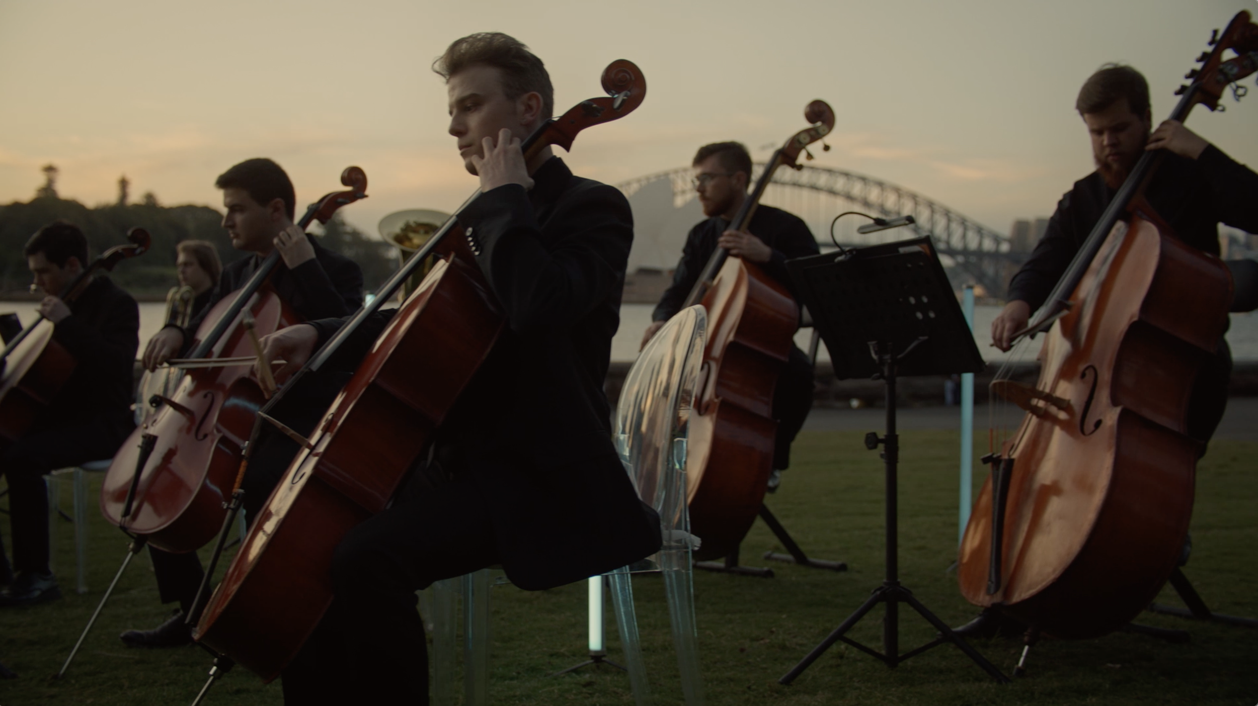 A group of musicians playing cellos outdoors during sunset, with a city skyline and a bridge in the background.
