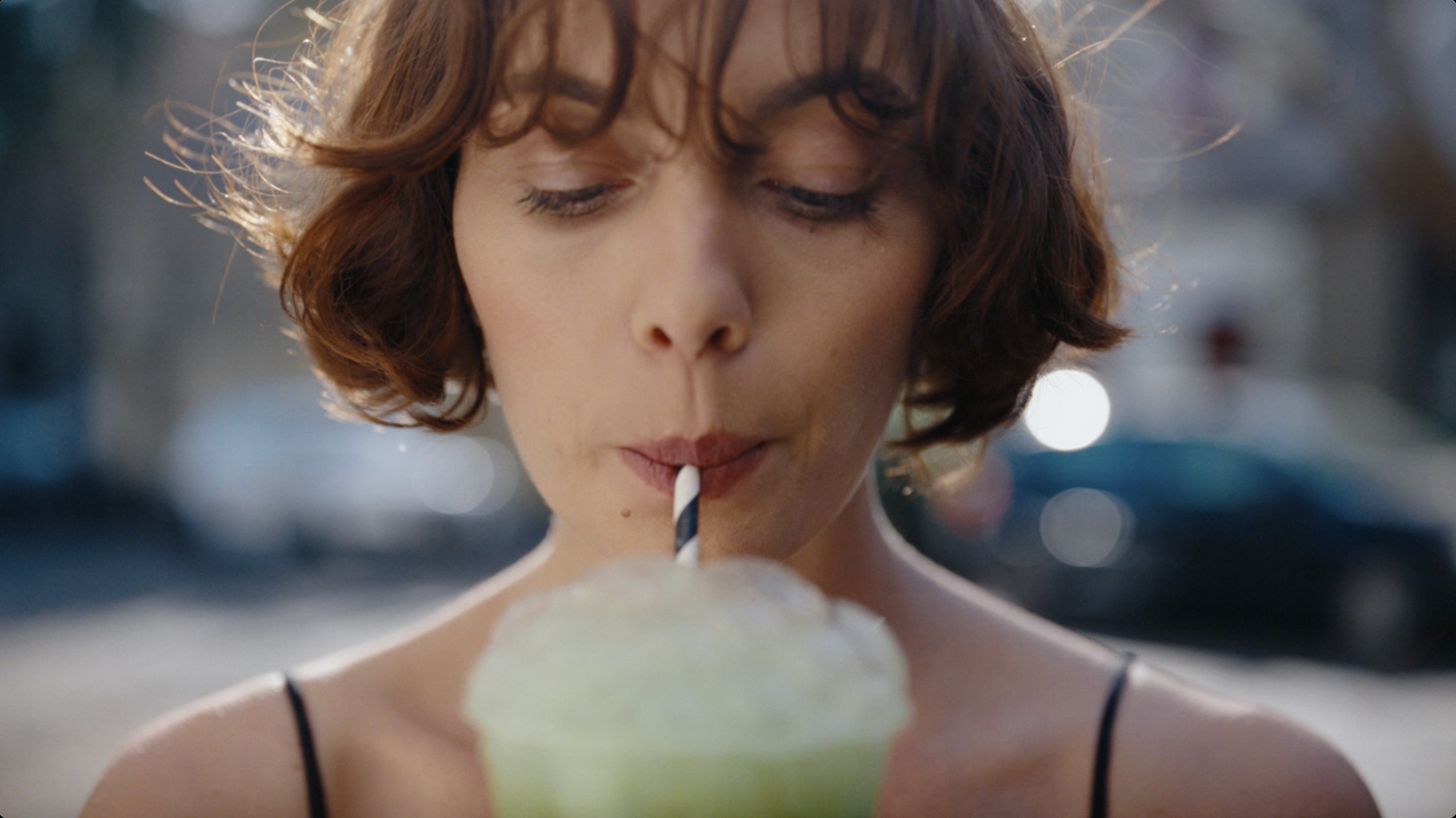 A woman with short wavy brown hair drinking a green frosty beverage through a striped straw.