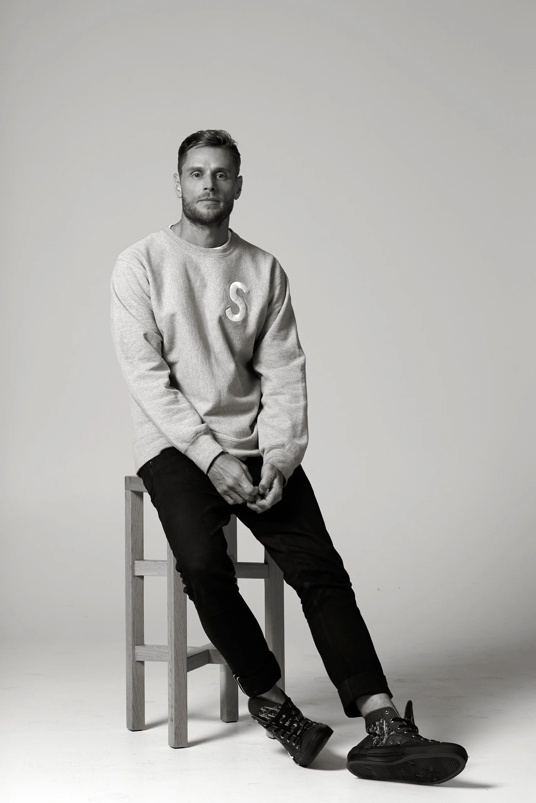 A young man with a beard and short hair, wearing a sweatshirt with the letter 'S', black jeans, and patterned sneakers, is sitting on a wooden stool against a plain background.
